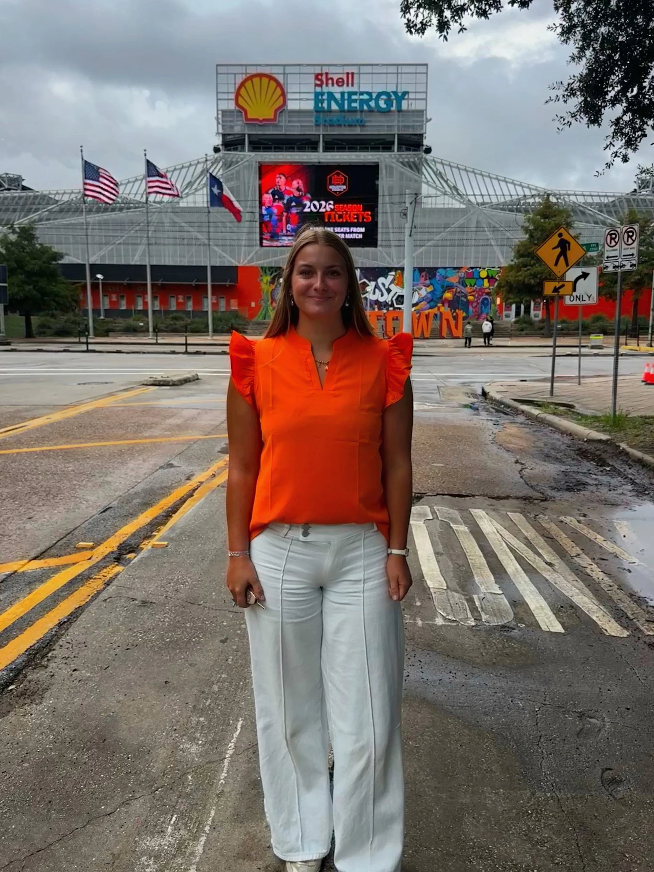 A woman wearing a bright orange top and white pants standing on a street with a stadium in the background, displaying signs for Shell Energy and a large digital screen, under cloudy skies.
