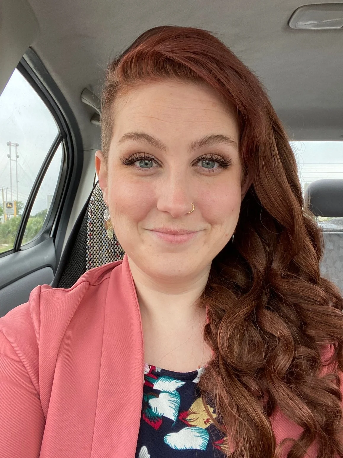 A photo of a white femme presenting person with freckles and blue eyes in a peach blazer with long red hair over one shoulder.