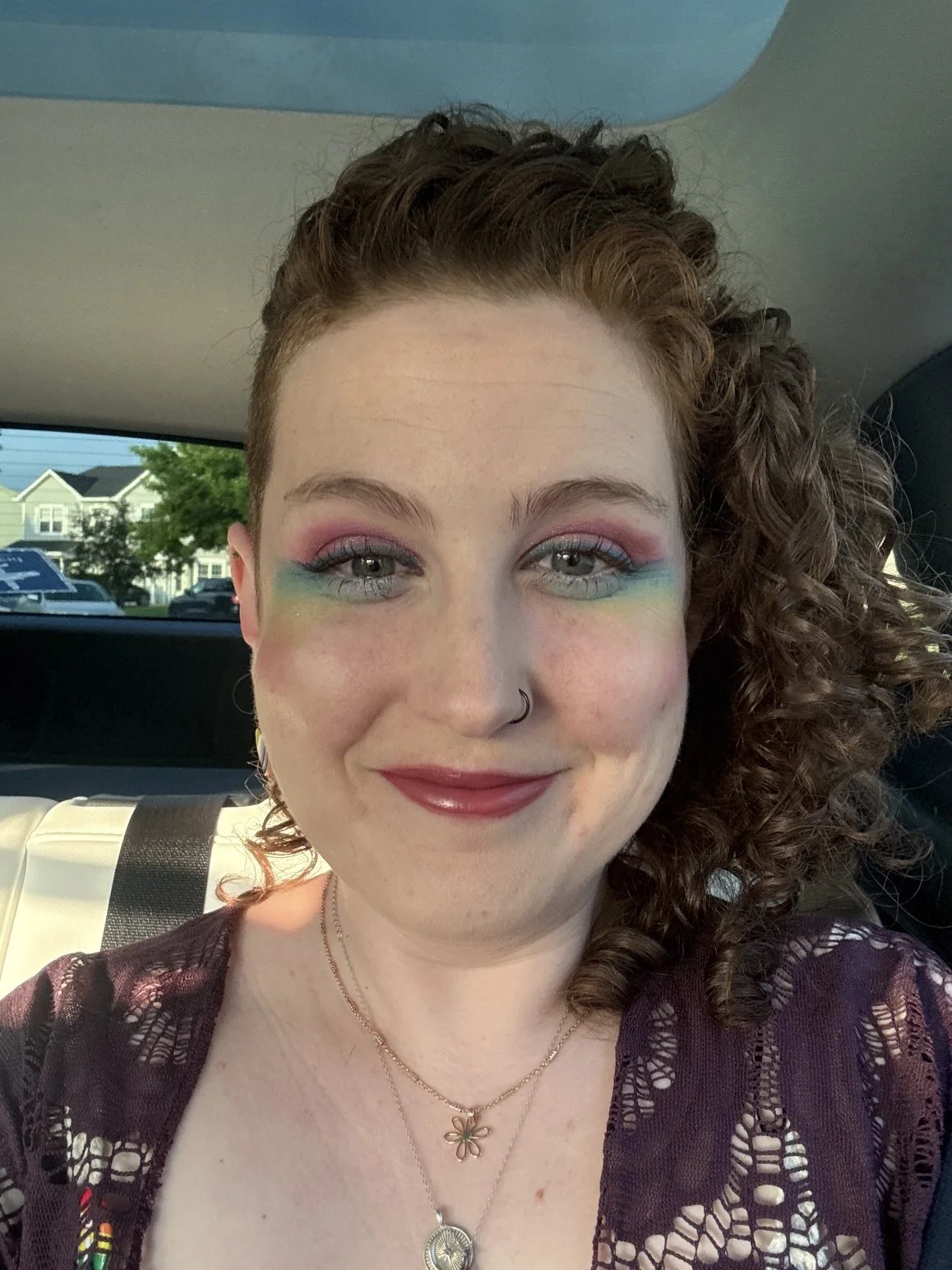 A white femme presenting person with freckles, curly auburn hair, and rainbow makeup around their eyes.