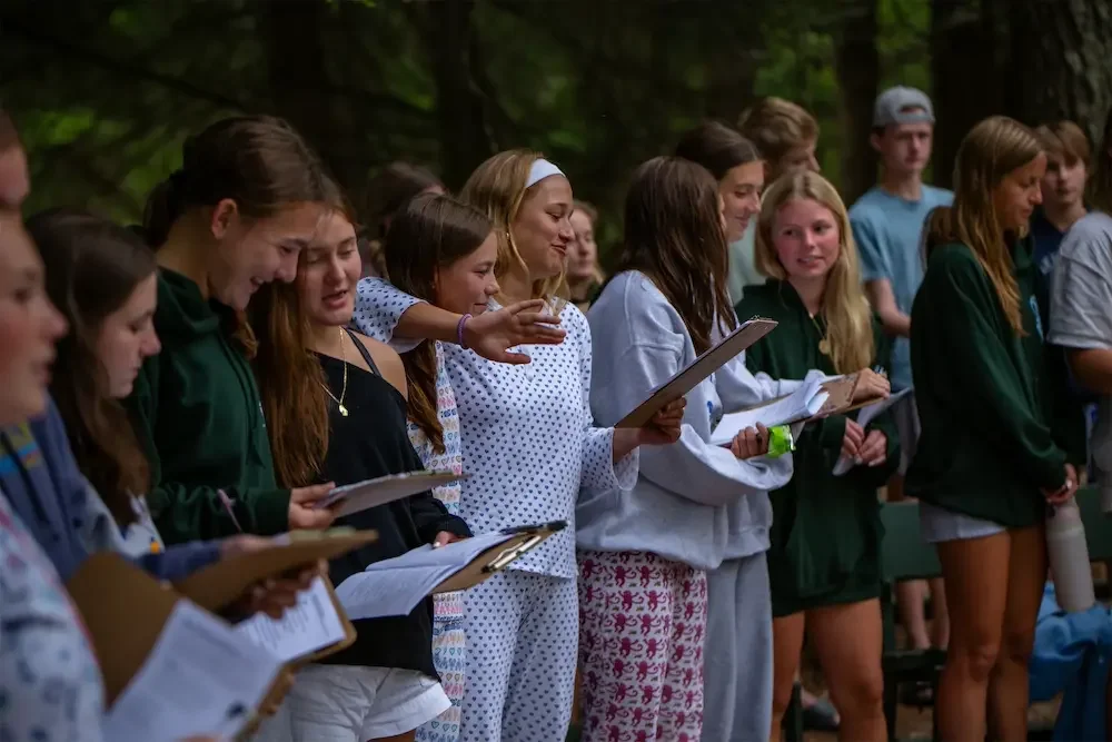 Group of young people outdoors, holding papers, likely participating in a reading or singing activity, with trees in the background.
