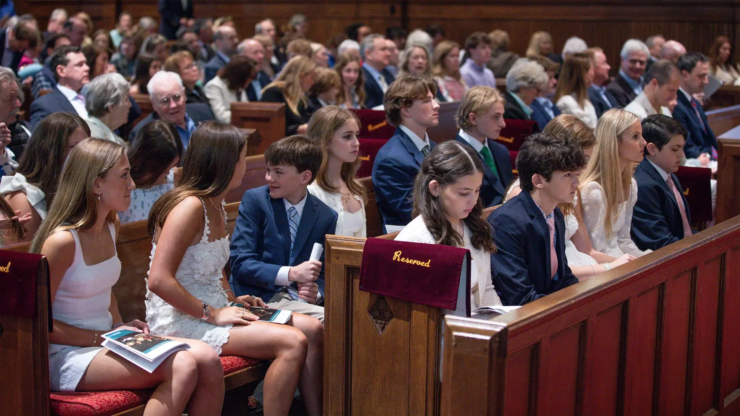 A group of people, mostly teenagers, dressed formally, sitting on church pews during a ceremony or event in a church or auditorium.