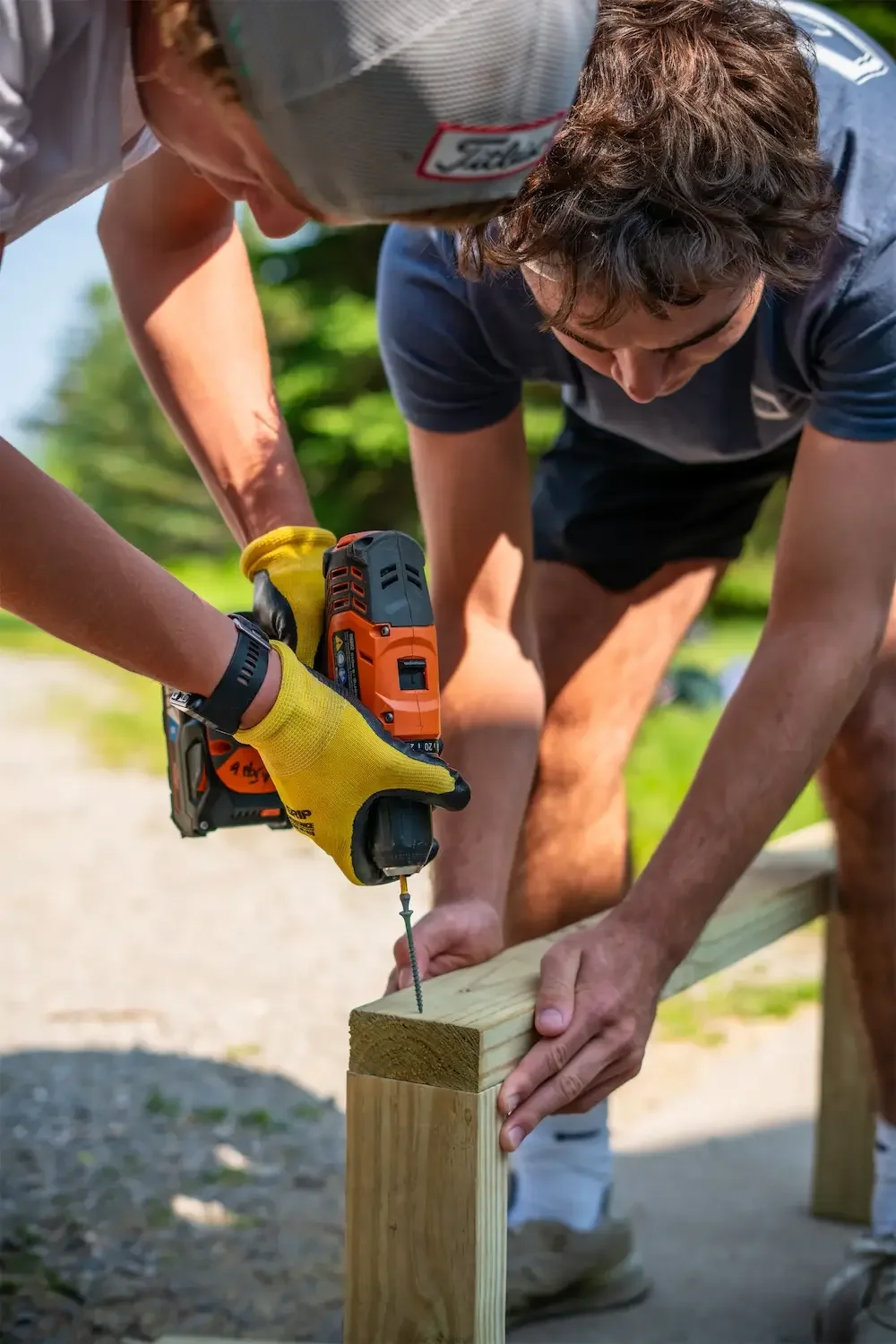 Two men working together outdoors, one using a cordless drill to screw into a wooden post while the other stabilizes it.