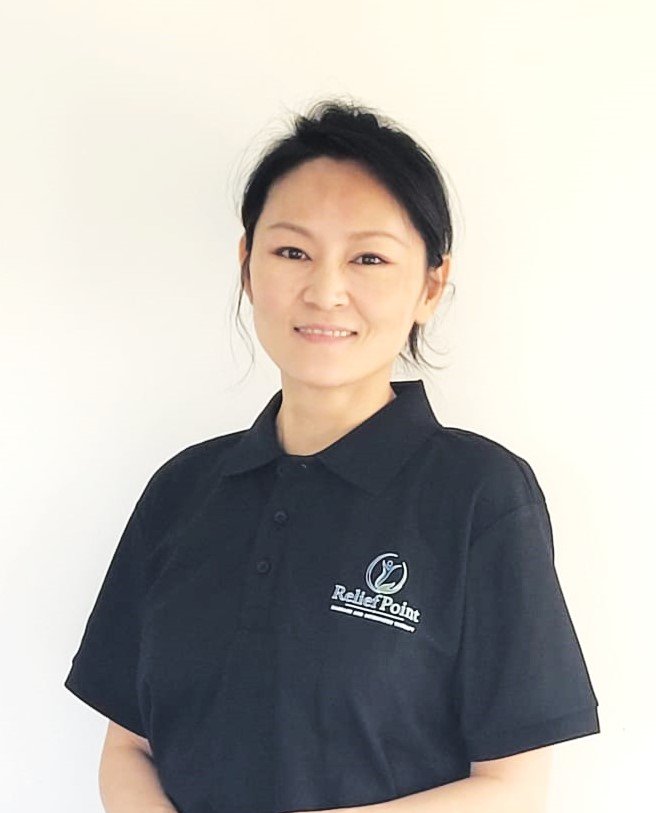 A woman with black hair in a ponytail, wearing a black polo shirt with a logo that reads 'Relief Point', standing against a plain white background.