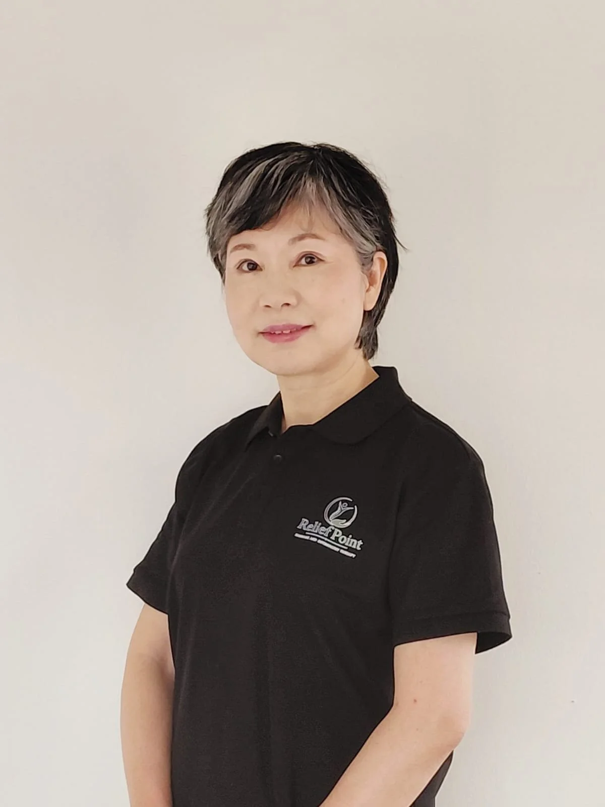 A woman with short black and gray hair wearing a black polo shirt standing against a plain white wall.
