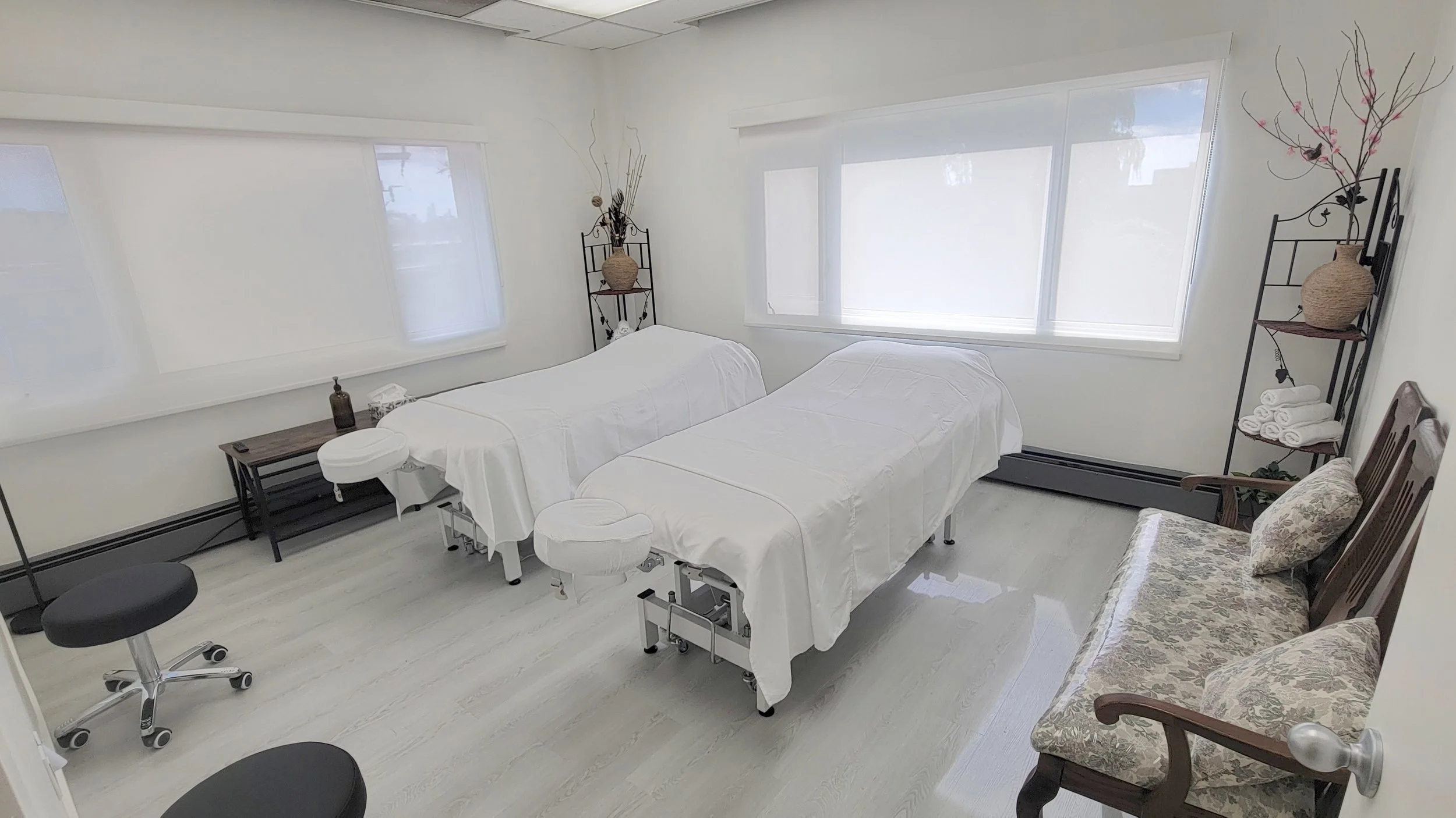 A room with two massage tables covered in white sheets, black shelving units with decorative vases and rolled towels, a floral upholstered chair, and large windows with white blinds letting in natural light.