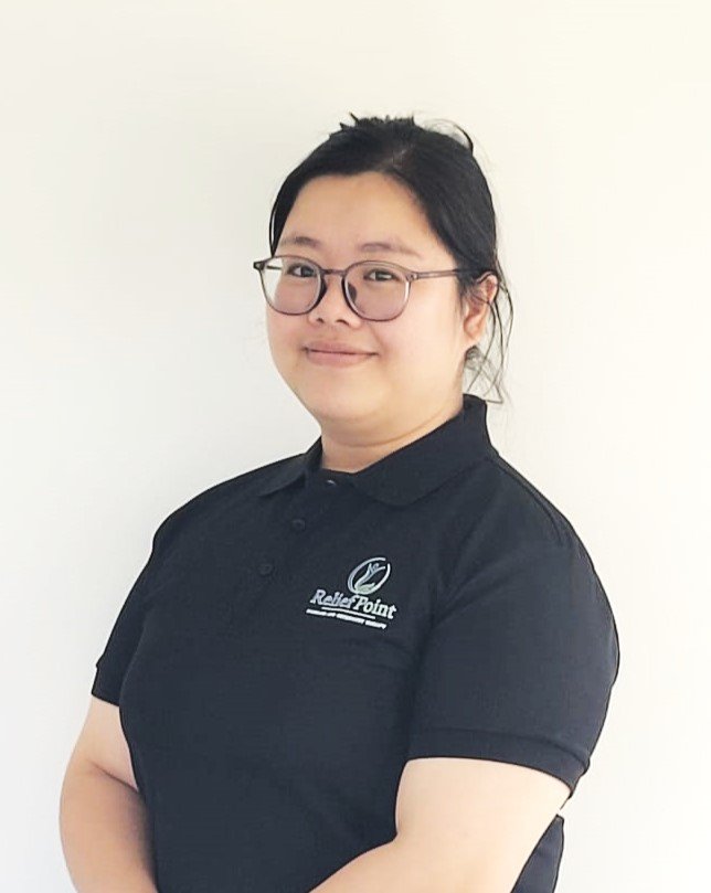 A woman with short black hair and glasses, wearing a black polo shirt with a Relief Point logo, standing against a plain white wall.