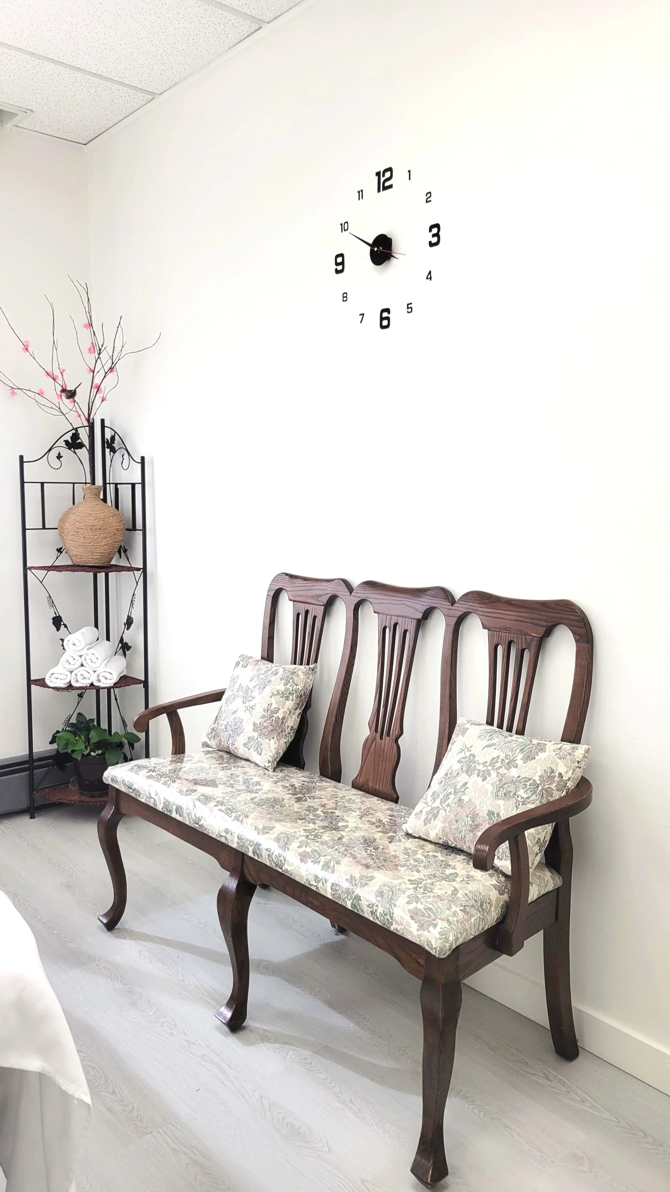 A waiting room with three wooden chairs with upholstered cushions and a floral pattern, a black metal shelving unit with a large vase, decorative branches with pink flowers, rolled white towels, and a small potted plant. A large wall clock with black numbers and hands is on the white wall.