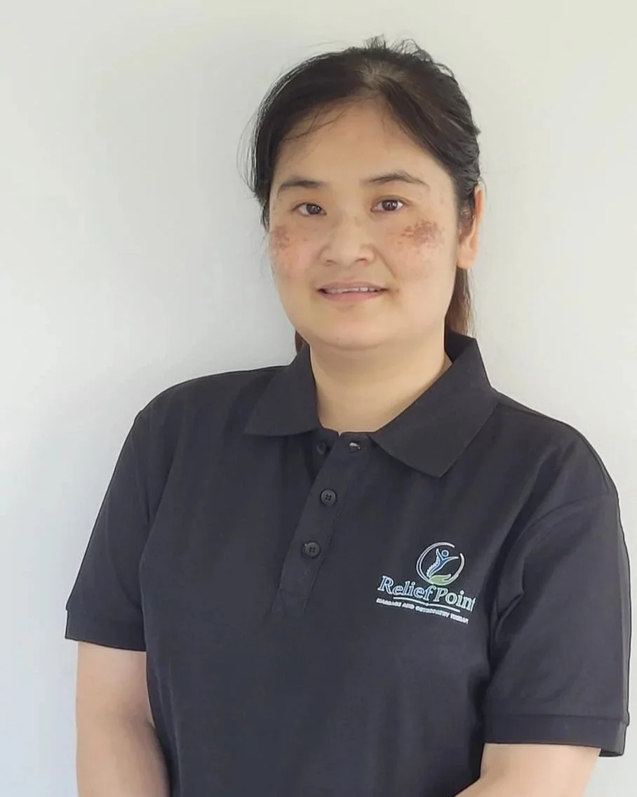 A woman with dark hair tied back, wearing a black polo shirt with a logo that says "Relief Point" on the left side, standing against a white background.