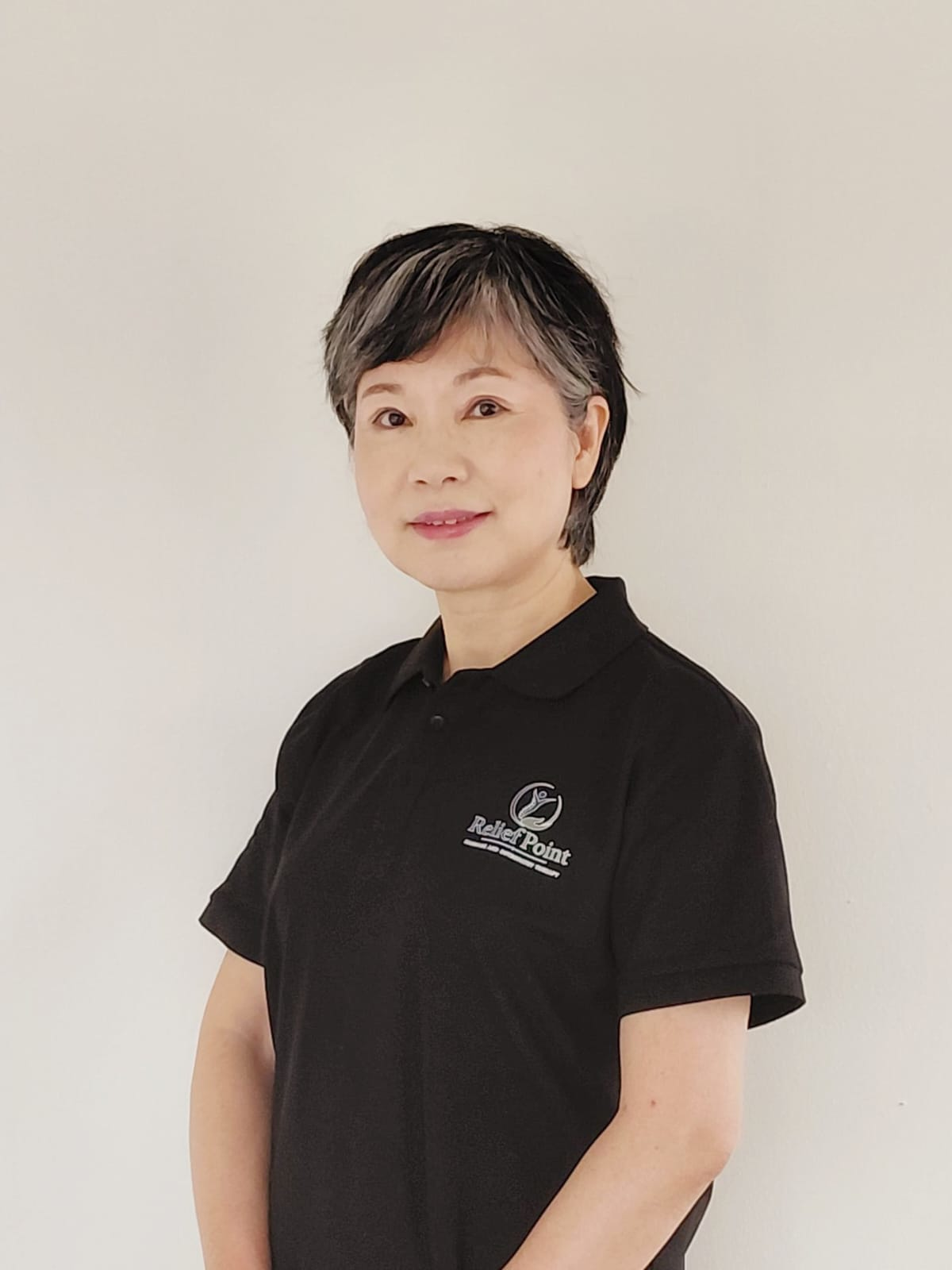 A woman with short black hair, wearing a black polo shirt with a logo that says 'Relief Point' on the chest, standing against a plain white wall.