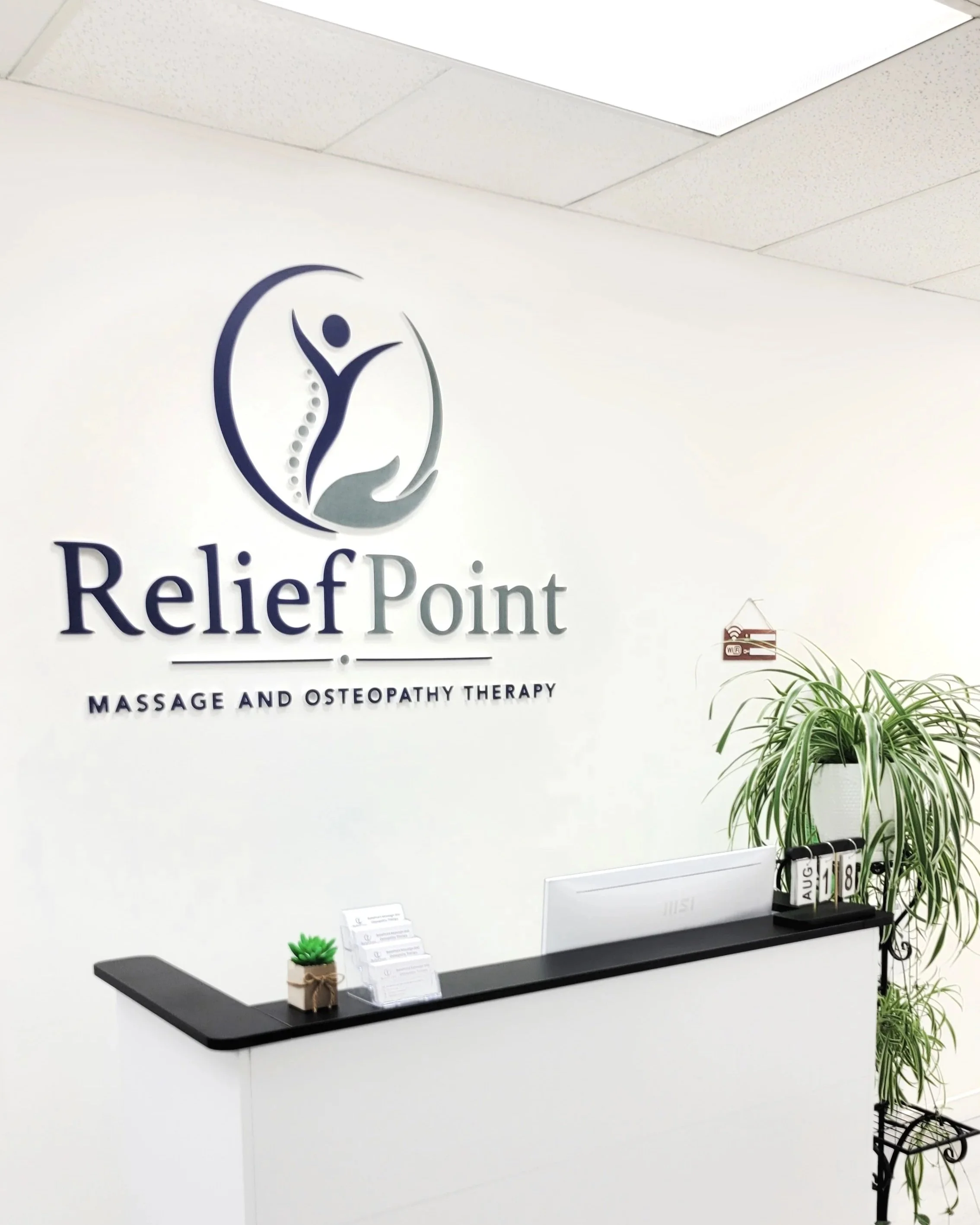 Reception area of a massage and osteopathy therapy clinic called ReliefPoint, featuring a black table with a small potted plant, informational flyers, a flip calendar, potted plants, and a sign with the clinic's name and logo on a white wall.