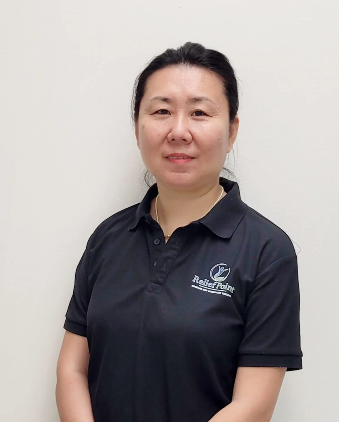 A woman with dark hair wearing a black polo shirt with a 'ReliefPoint' logo, standing against a plain white wall.