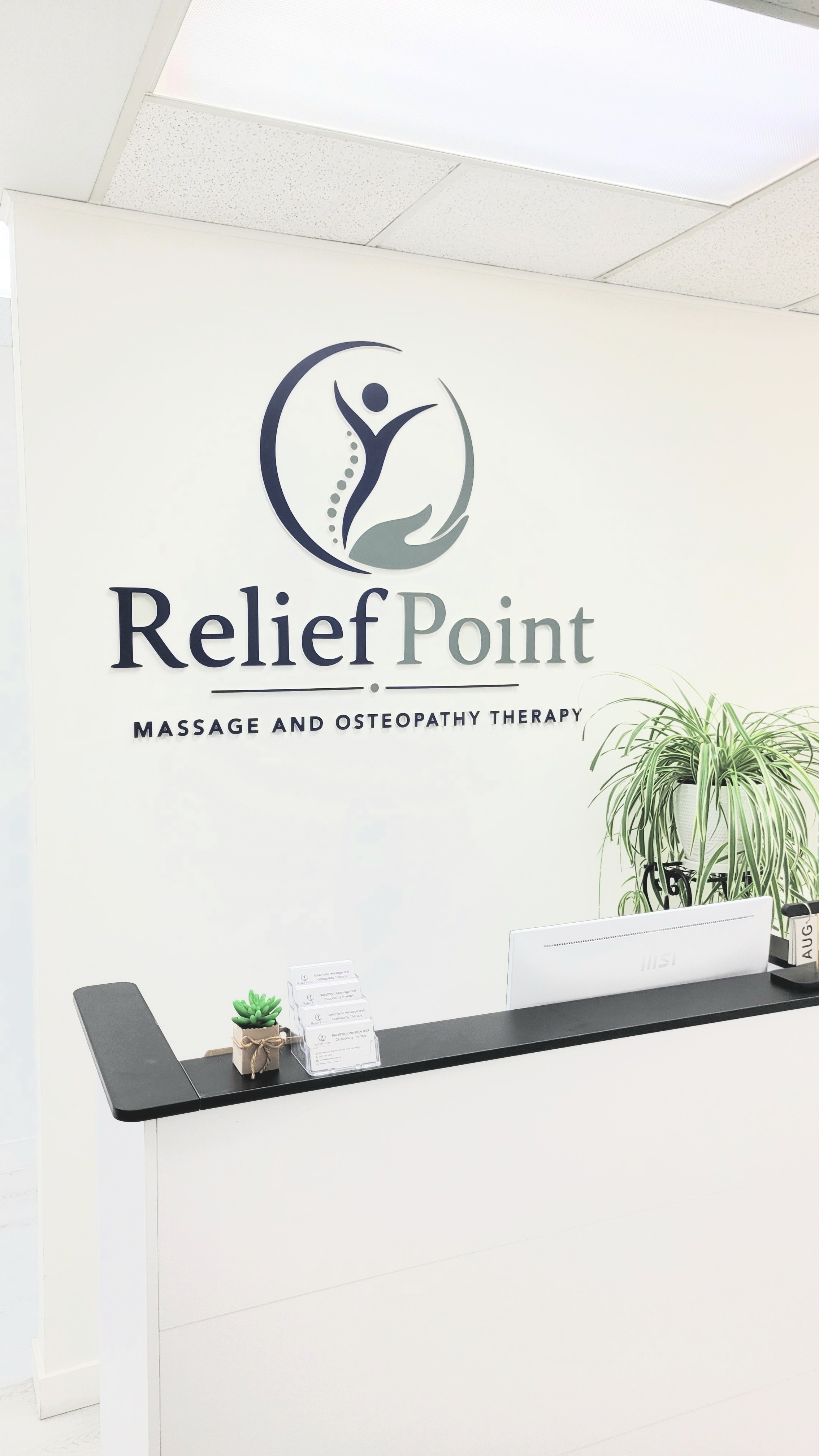 Reception desk with a small plant, pamphlets, a computer, and a sign that reads 'Relief Point Massage and Osteopathy Therapy' on the wall behind, along with a tall plant to the right.
