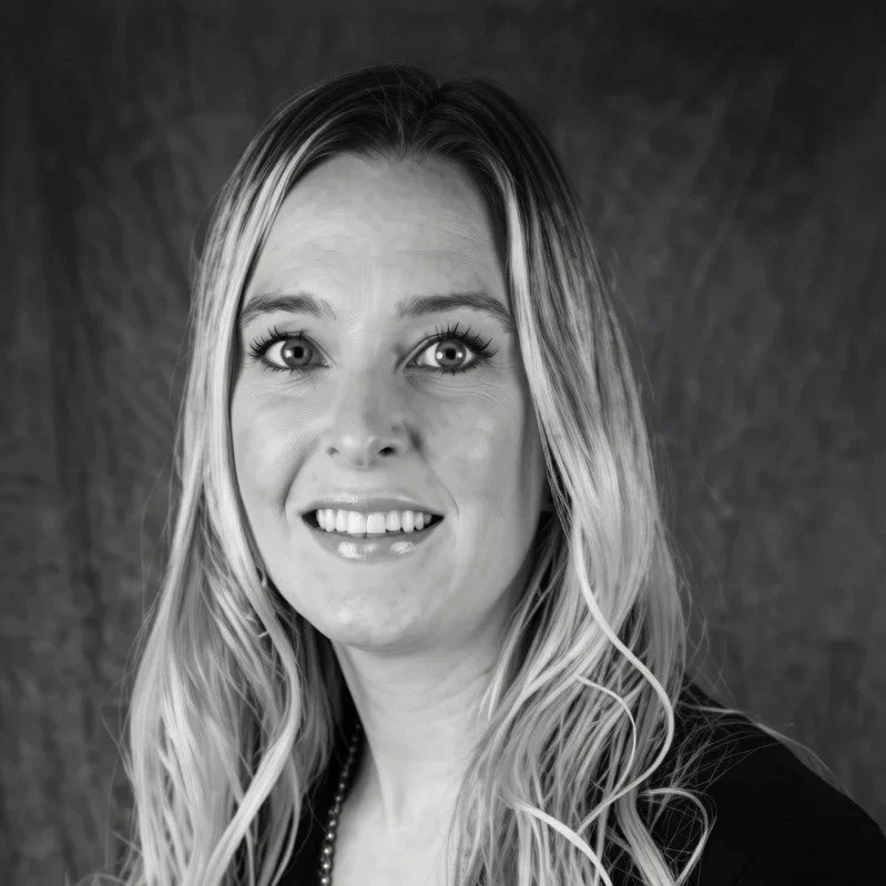 Black and white portrait of a woman with long, wavy hair, smiling, wearing a dark top and a beaded necklace against a textured background.