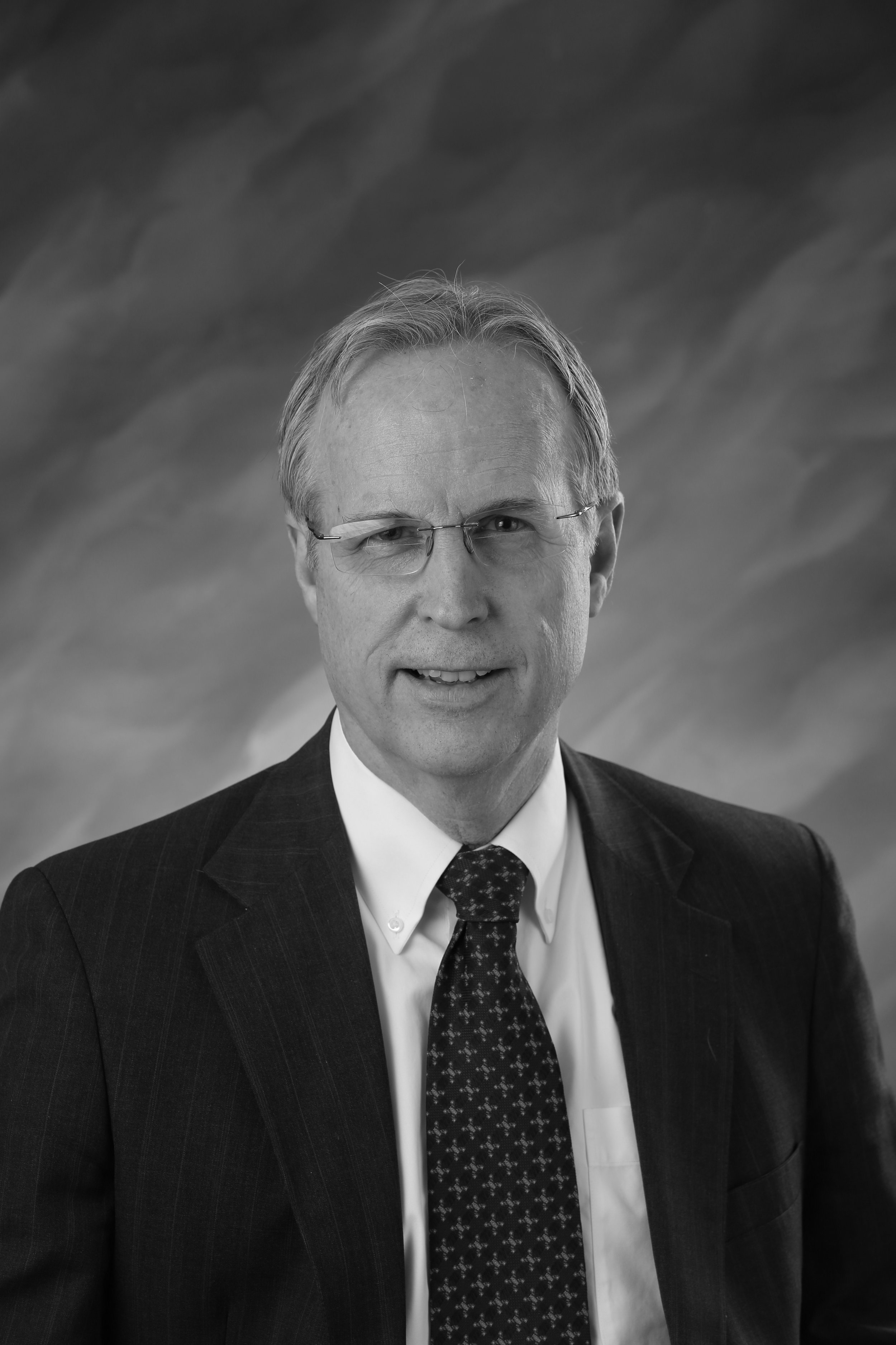 Black and white portrait of a middle-aged man wearing glasses, a dark pinstripe suit, a white shirt, and a patterned tie, against a cloudy sky background.