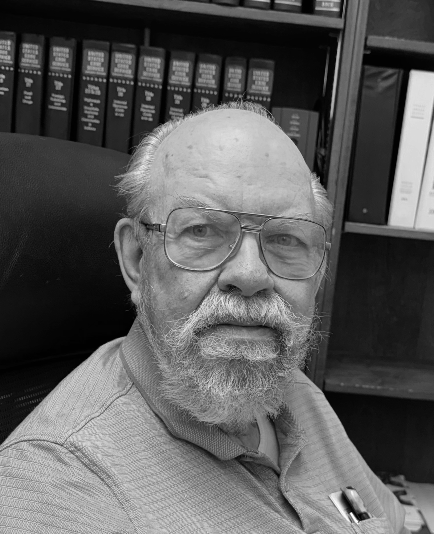 A black and white photo of an older man with a bald head, glasses, and a beard, sitting in front of a bookshelf filled with large reference books, wearing a collared shirt with a pen in the pocket.