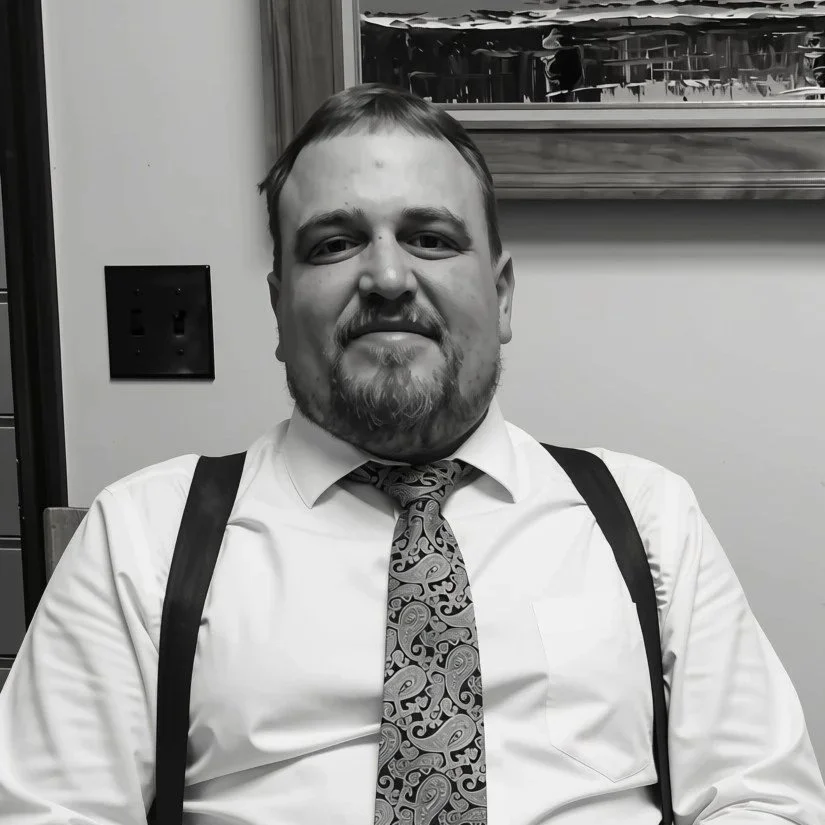 A man with short hair, a beard, and a mustache, dressed in a white shirt, patterned tie, and suspenders, sitting indoors with a framed picture on the wall behind him.