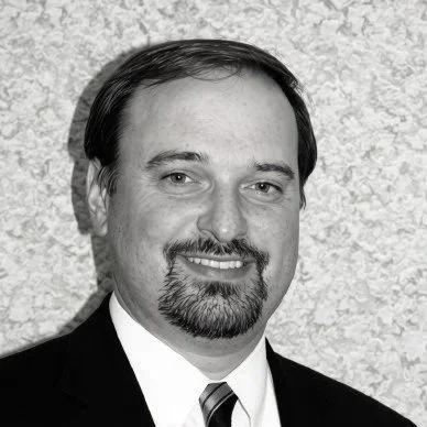 A black-and-white headshot of a man with dark hair, a goatee, wearing a suit and tie, smiling at the camera.