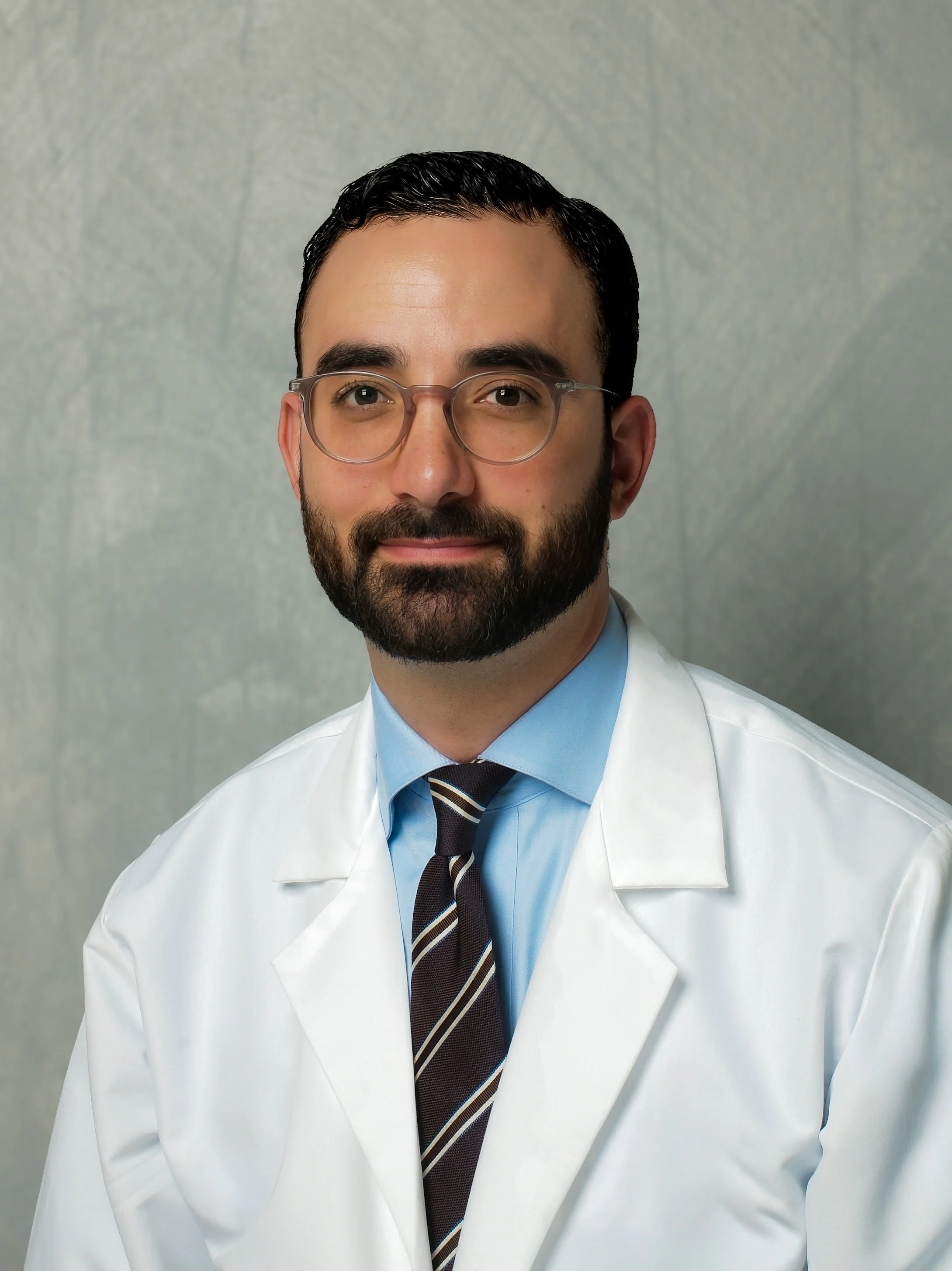 A man with glasses and a beard wearing a white lab coat, light blue shirt, and tie, standing against a neutral background.