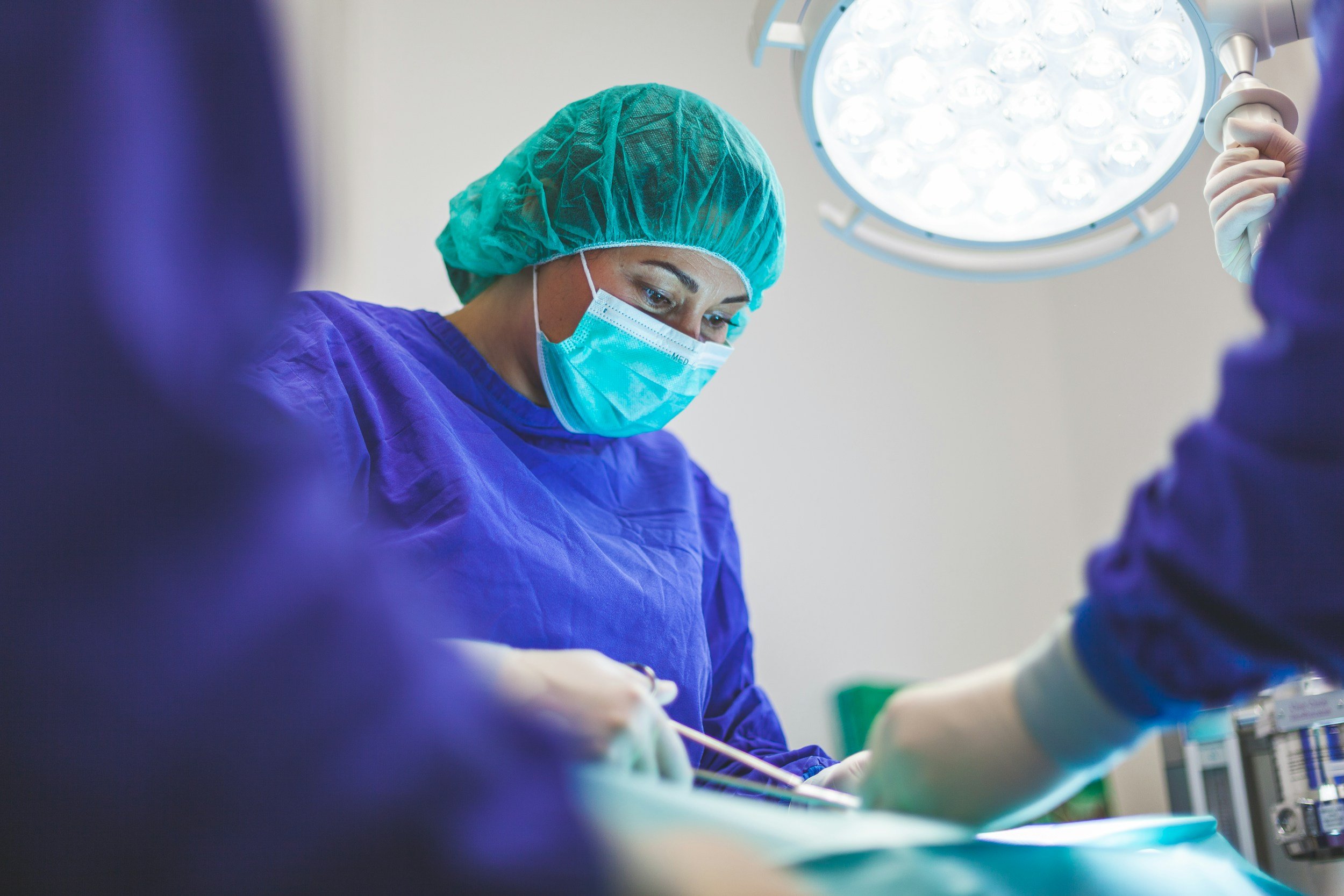 A female surgeon wearing a surgical mask, green hair covering, and purple gown performing surgery in an operating room, illuminated by a bright overhead light.