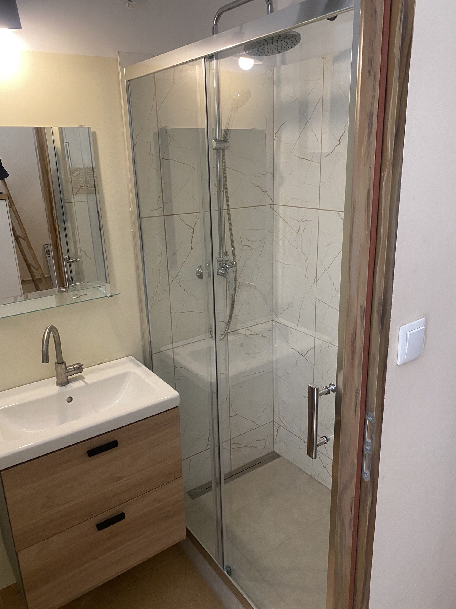 A modern bathroom featuring a glass shower enclosure with marble-patterned tiles, a wooden vanity with a white sink, and a wall-mounted mirror.