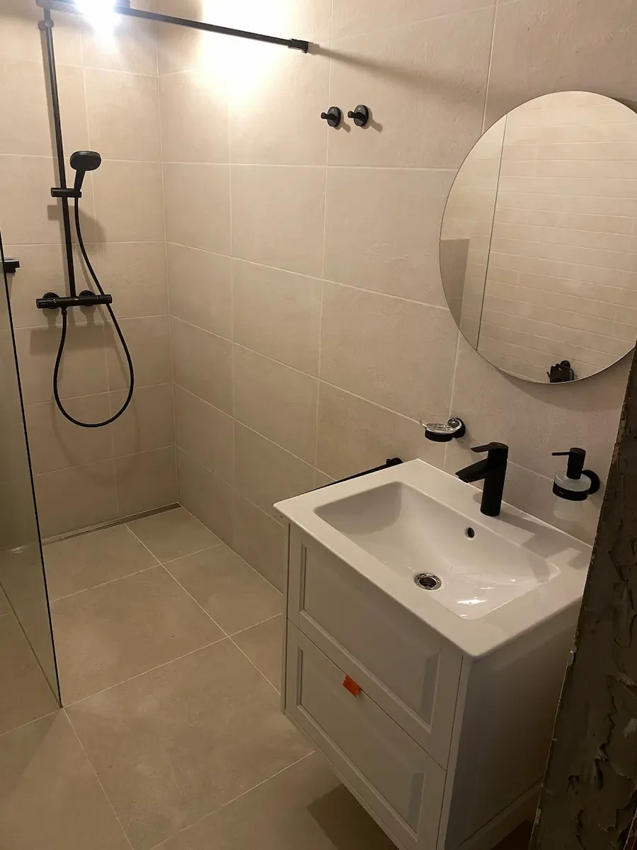 Nowoczesna łazienka z prysznicem typu walk-in, czarną deszczownicą i armaturą, białą umywalką z czarną baterią, okrągłym lustrem oraz beżowymi płytkami ściennymi i podłogowymi.