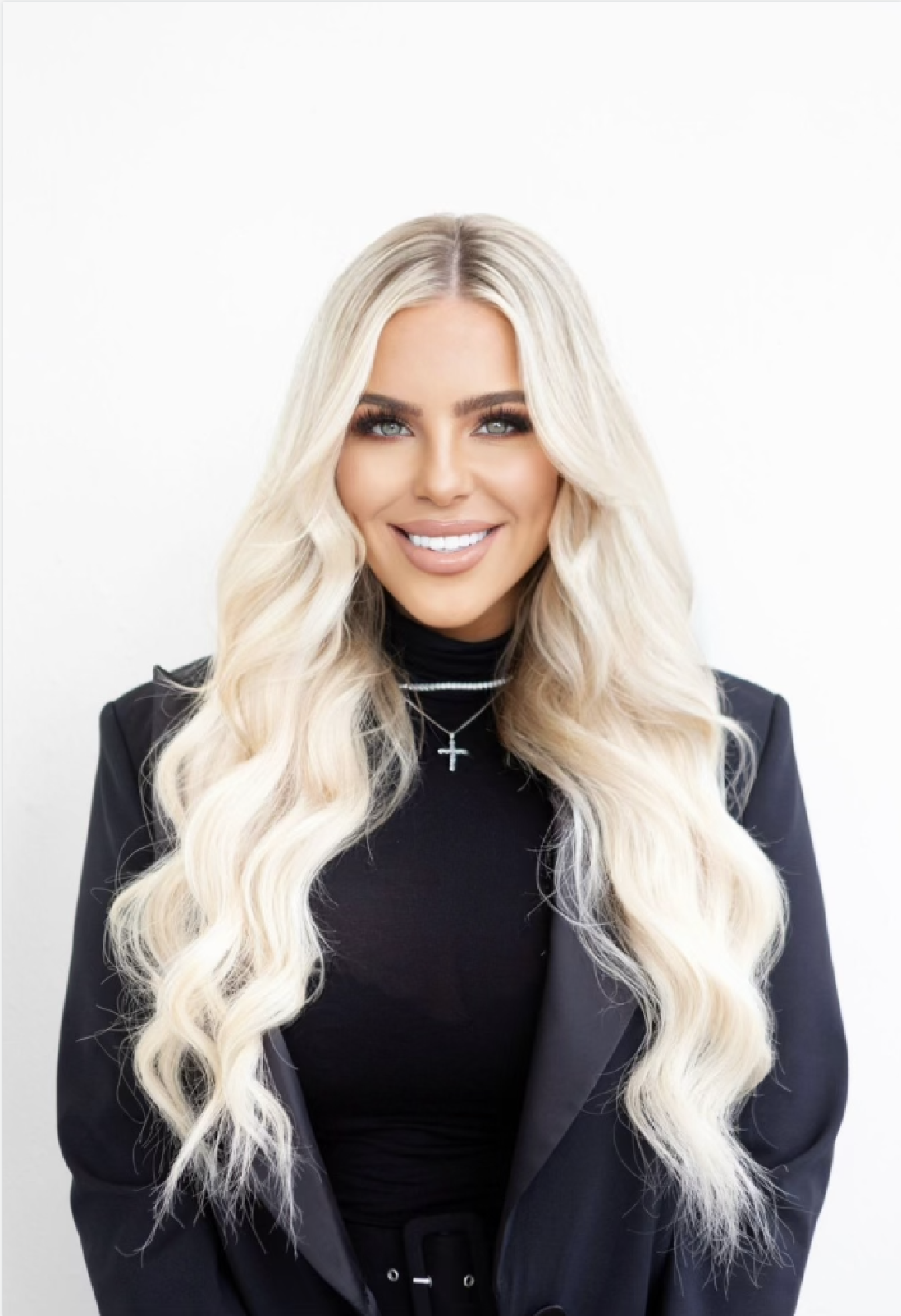 Portrait of a woman with long, wavy blonde hair, wearing a black blazer and a black top, smiling against a plain white background.