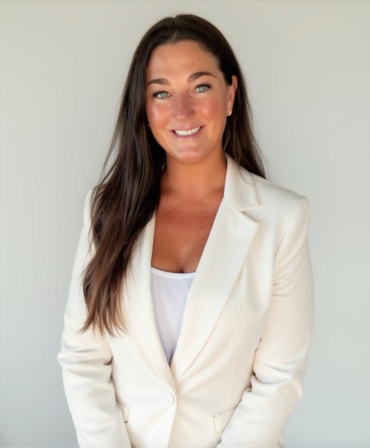 A smiling woman with long brown hair, blue eyes, and earrings, wearing a white blazer and white top, standing against a light gray background.