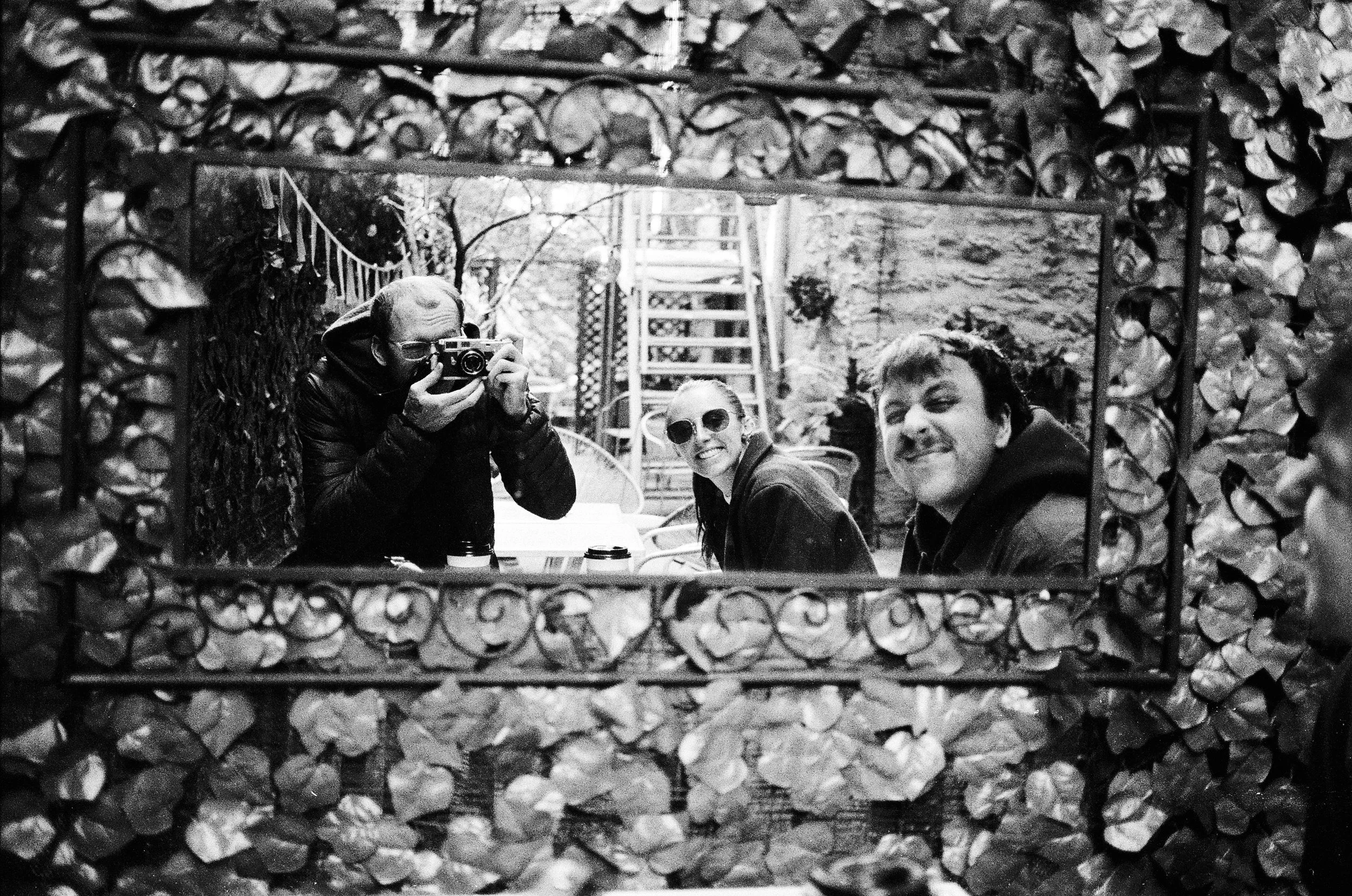 Group of three people taking a photo of each other through a decorative mirror with a woodpile background.