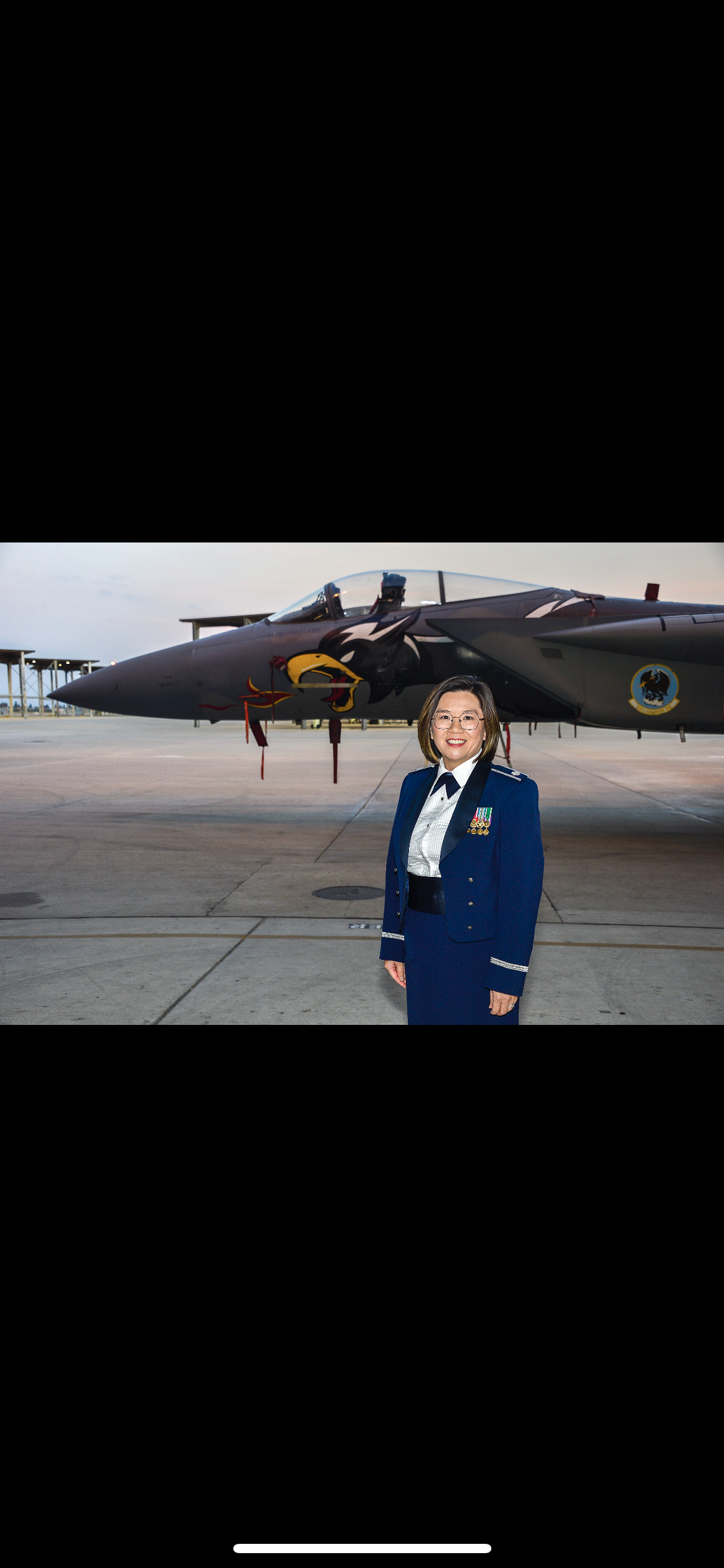 A woman in a military uniform standing in front of a jet aircraft at an airbase with a sunset sky in the background.