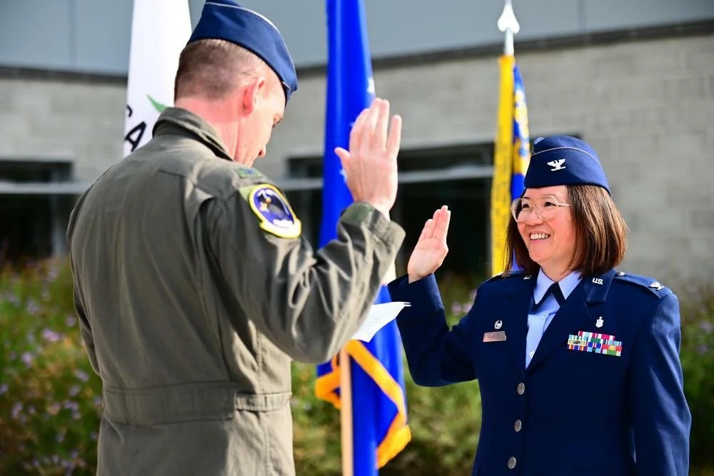 A woman in a blue military uniform and a woman in a beige military uniform are giving each other a high five outdoors, with United States flags in the background.