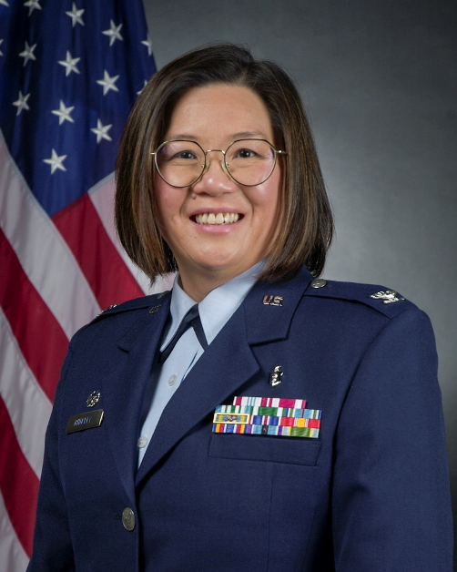 Portrait of a smiling woman in a U.S. Air Force uniform with medals and ribbons, standing in front of an American flag.