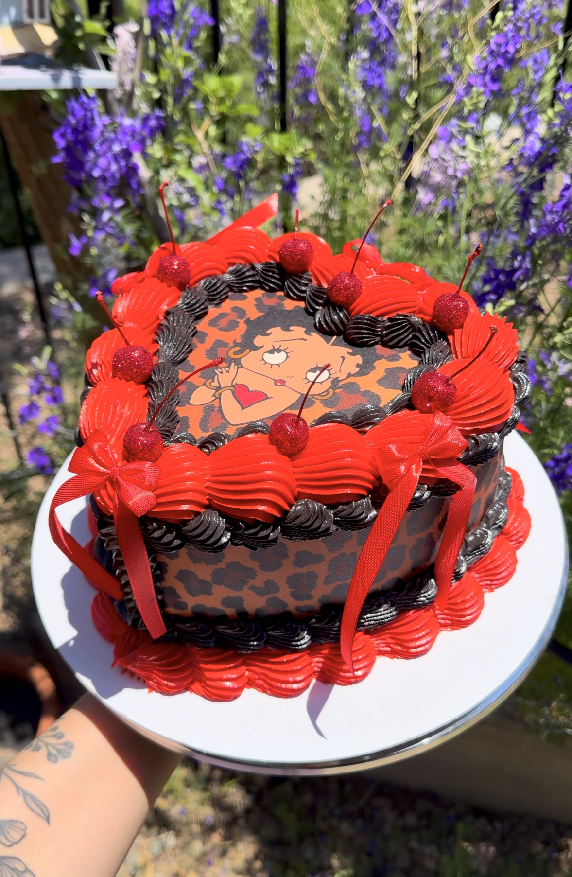 A cake decorated with red and black frosting, featuring a Betty Boop cartoon image on top, surrounded by red cherries and red ribbons, with purple flowers in the background.