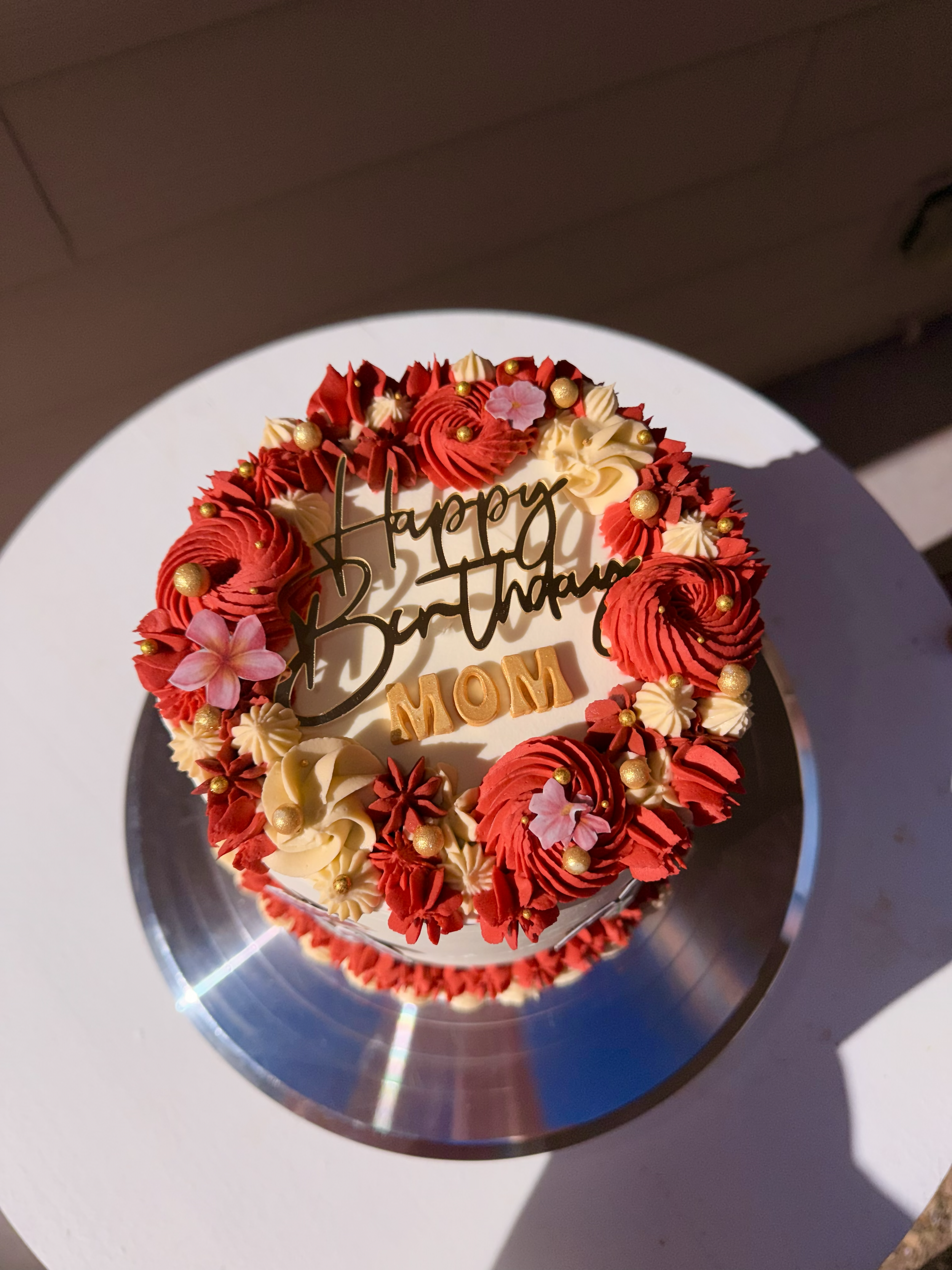 A round birthday cake decorated with red and white frosting, gold and pink edible beads, pink flowers, and a golden inscription that reads 'Happy Birthday MOM'. The cake is placed on a reflective silver cake board.