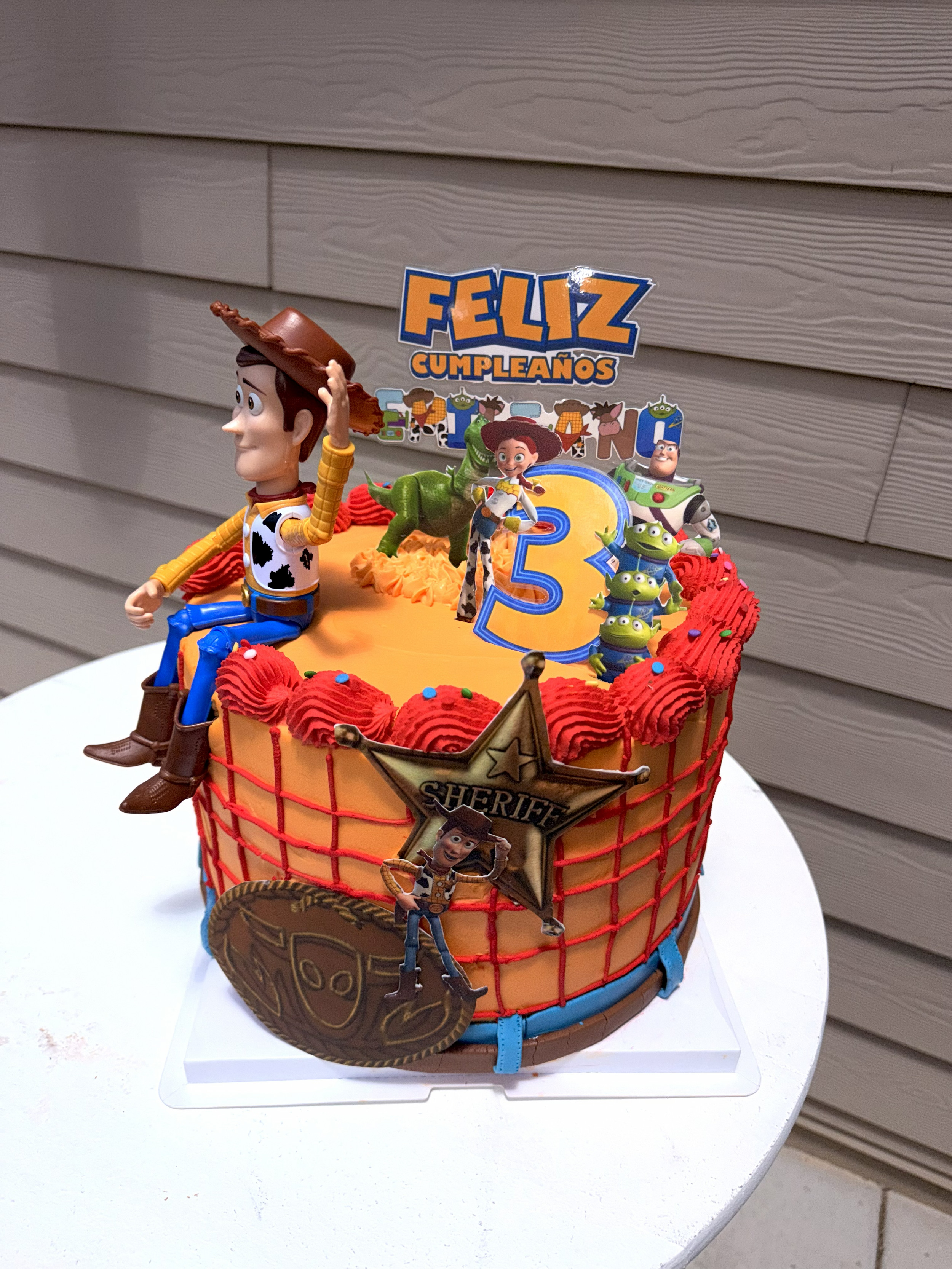 A Toy Story-themed birthday cake with figurines of Woody, Jessie, Rex, Buzz Lightyear, and characters from Toy Story, decorated with red icing, a sheriff's badge, and a large number 5 topper. The cake also has a sign that says 'Feliz Cumpleaños'.