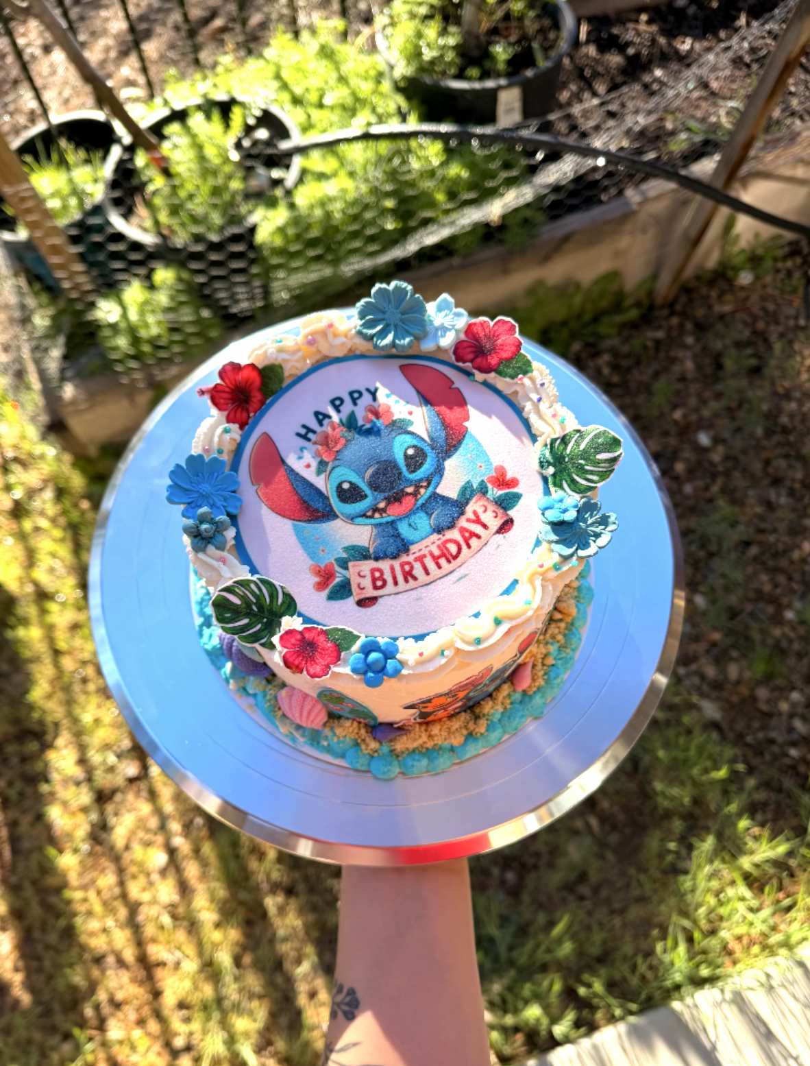 Colorful birthday cake with a Stitch character edible topper, decorated with blue and red flowers, green leaves, and pink details, held outdoors with sunlight.