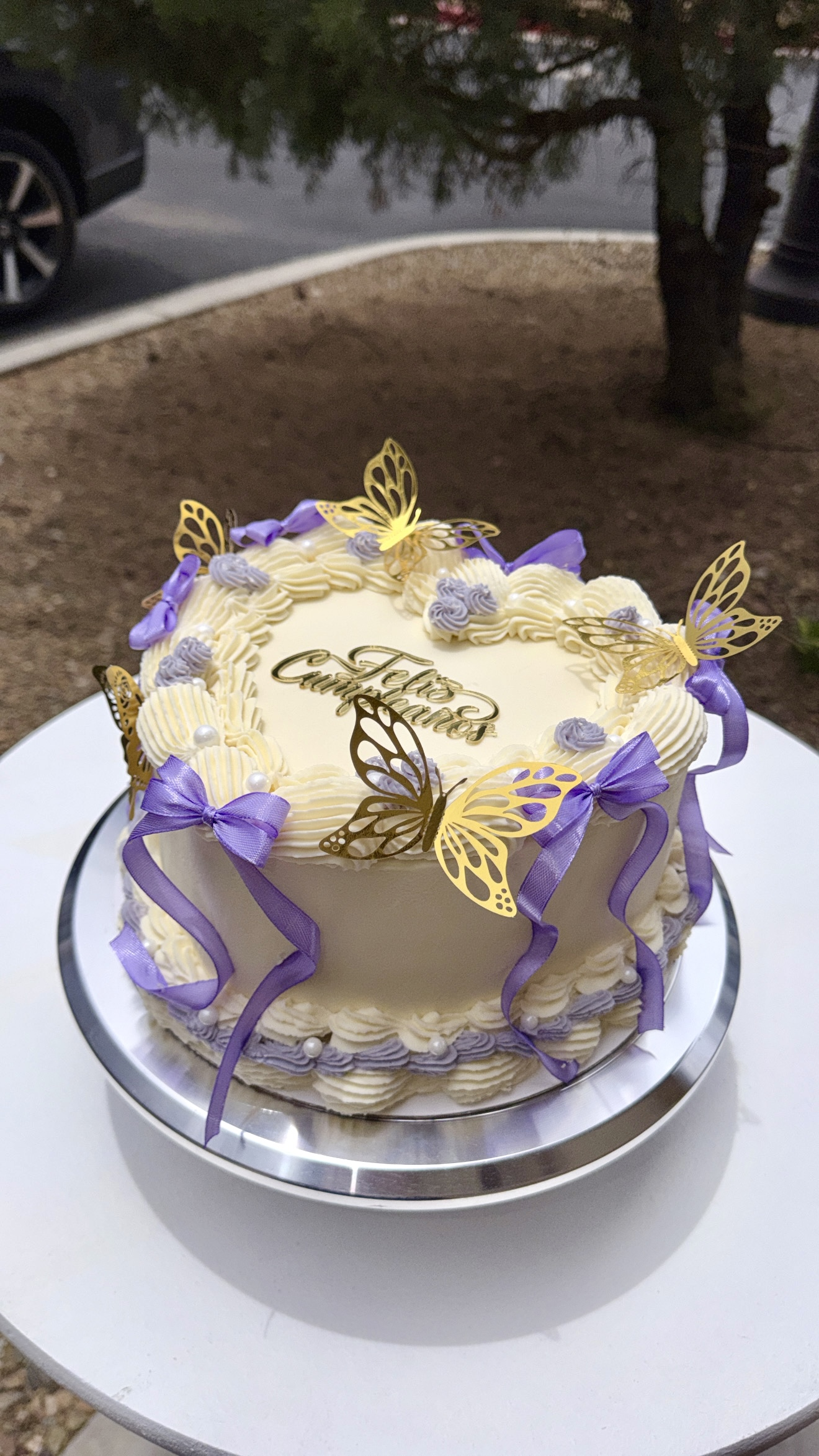 A cream-colored birthday cake decorated with purple ribbons, yellow butterfly cutouts, and purple frosting roses. The cake has a golden "Happy Birthday" topper and is placed on a silver cake stand.