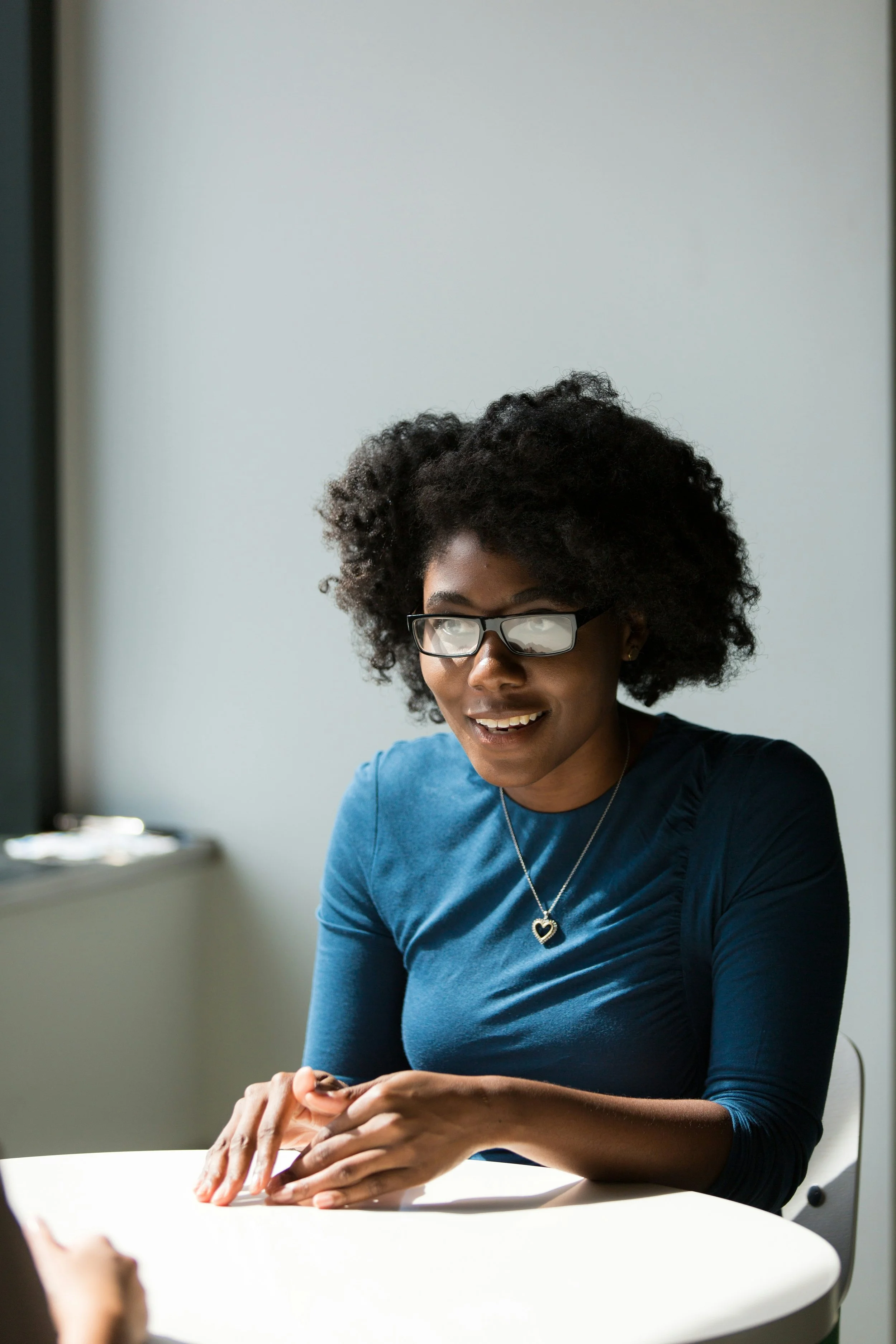 A young woman with curly hair and glasses, wearing a blue shirt and heart-shaped necklace, smiling and looking down at her hands, sitting at a white table with a plain wall in the background.