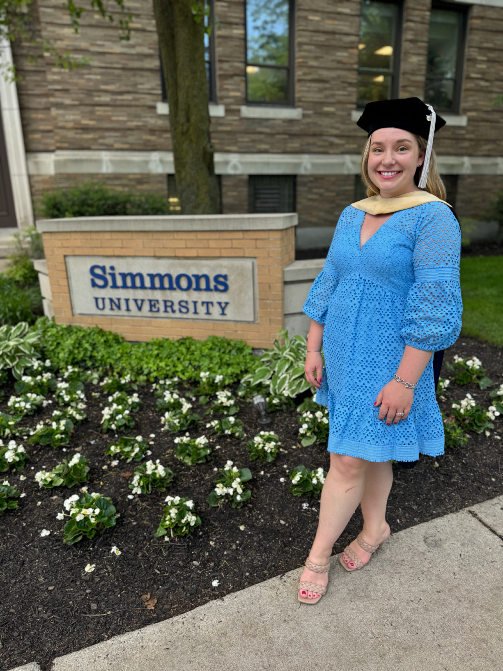 A woman in a blue dress and beige heels standing outside Simmons University, wearing a graduation cap and gown. There are white flowers in the garden bed and a brick building in the background.
