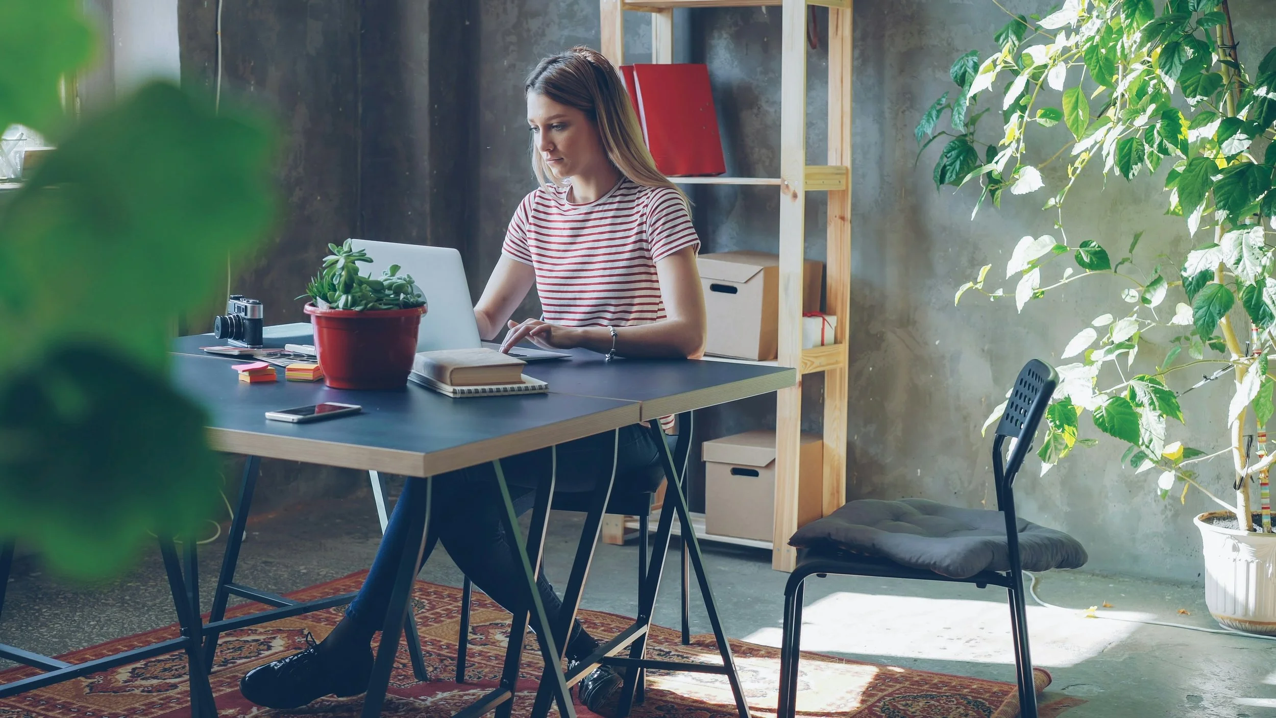 A woman sitting at a desk working on a laptop in a modern, industrial-style office with plants and shelves.