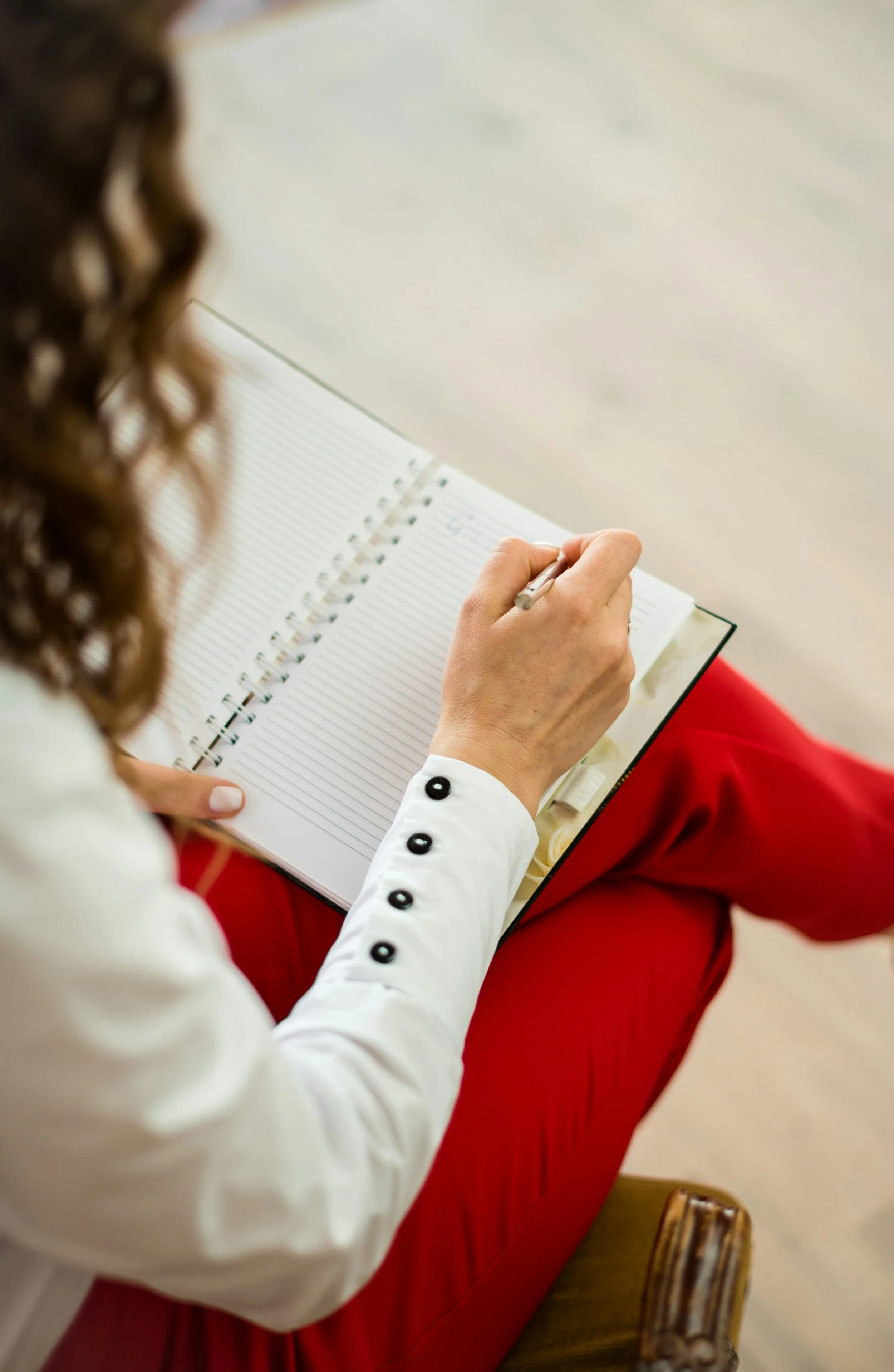 Person sitting on a red chair, writing in a notebook with lined pages. The person is wearing a white long-sleeved shirt with black buttons on the sleeve and red pants. The image is taken from behind the person, with curly brown hair visible on the left.