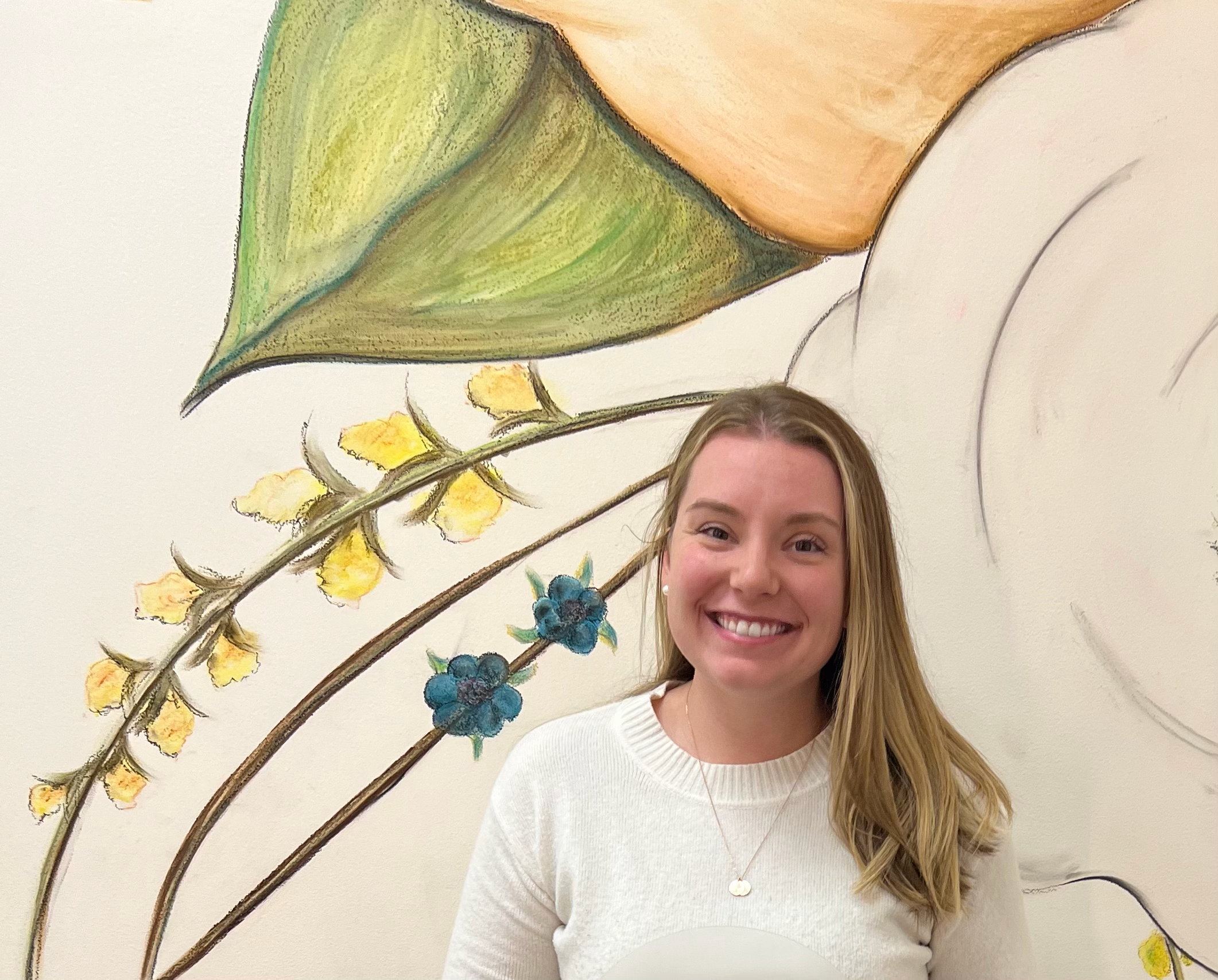 A woman with blonde hair smiling in front of a wall with a colorful botanical mural featuring green leaves, yellow and blue flowers.