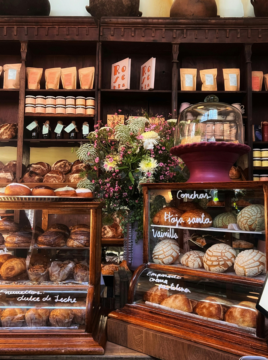 Colorful French style bakery with breads and shelves of goods. There's a large bouquet of flowers in the center. The image is warm and rustic.