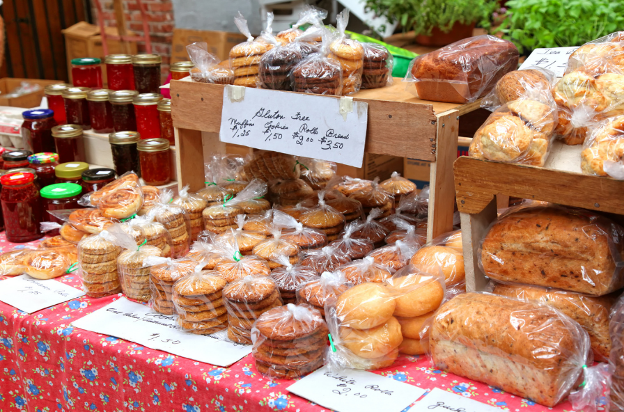 Baked goods and jams on display at an outdoor market stall, including cookies, bread, and muffins wrapped in plastic bags, with handwritten price signs on white paper.