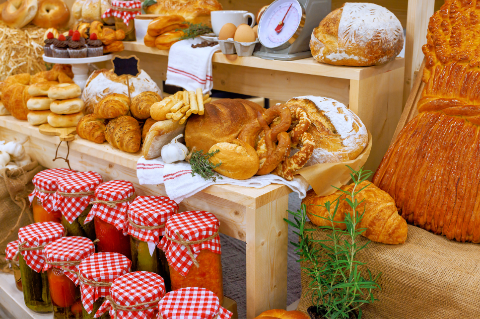 A display of baked goods including bread, croissants, pastries, muffins, and a loaf, with jars of preserved vegetables, a scale, and a plant, suggesting a market or bakery setting.
