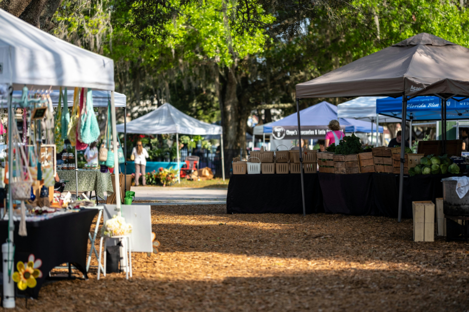 Outdoor farmer's market with vendor stalls selling crafts and produce on a sunny day amid trees.