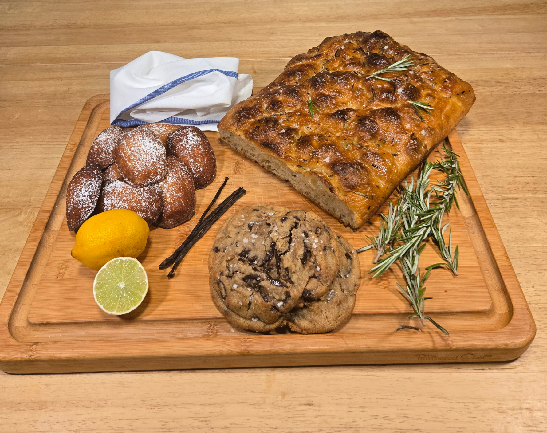 A wooden cutting board with lemon slices, a pile of cookies, a chocolate chip cookie, a large rectangular pizza topped with cheese and herbs, sprigs of rosemary, and a set of napkins.