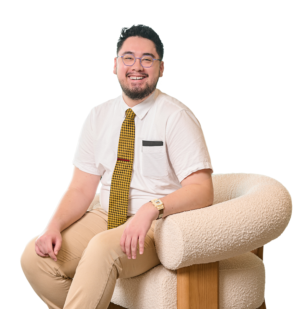 A smiling man with glasses and a beard wearing a white shirt, yellow checkered tie, beige pants, and a gold watch sitting on a beige textured armchair.
