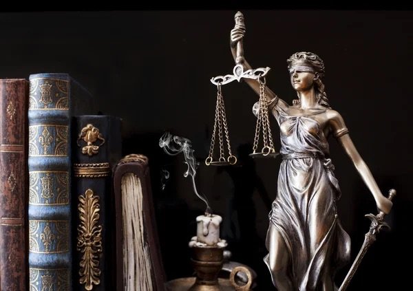 Decorative statue of Lady Justice holding scales and a sword, surrounded by antique books, a lit candle, and a quill on a dark background.