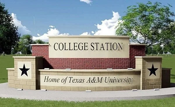 Sign reading "College Station, Home of Texas A&M University" with brick and stone design, black stars, trees, grass, and a sky with clouds in the background.