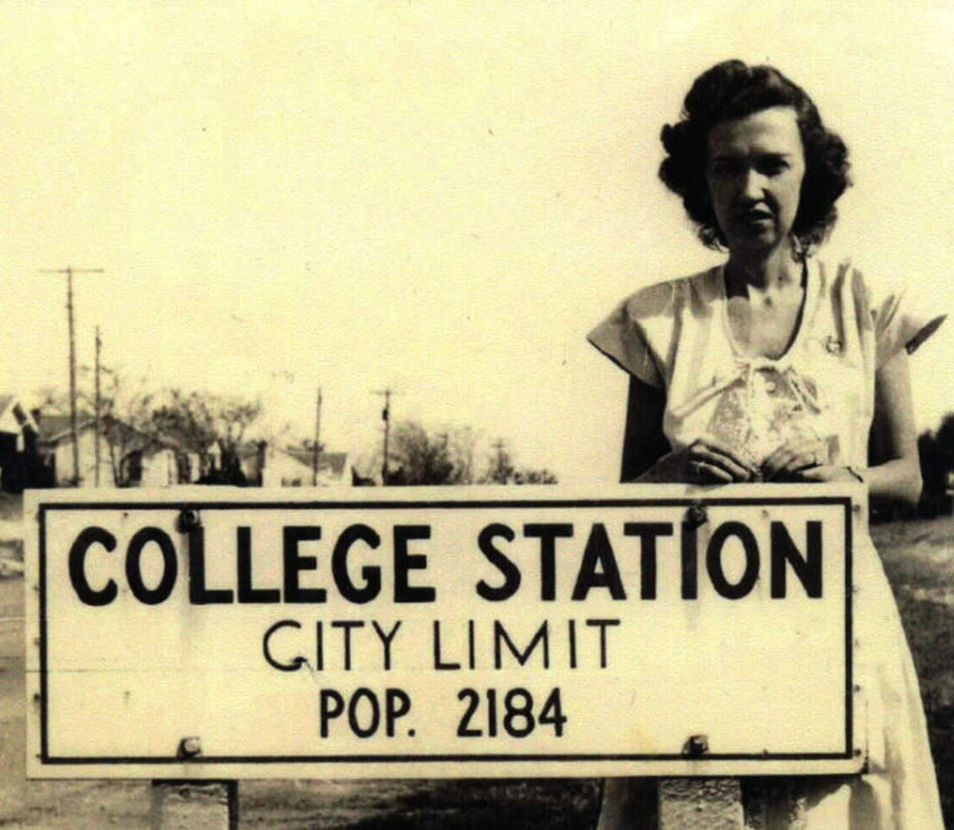 Black and white photo of a woman standing outdoors next to a sign that reads 'College Station City Limit Pop. 2184', likely from the mid-20th century.